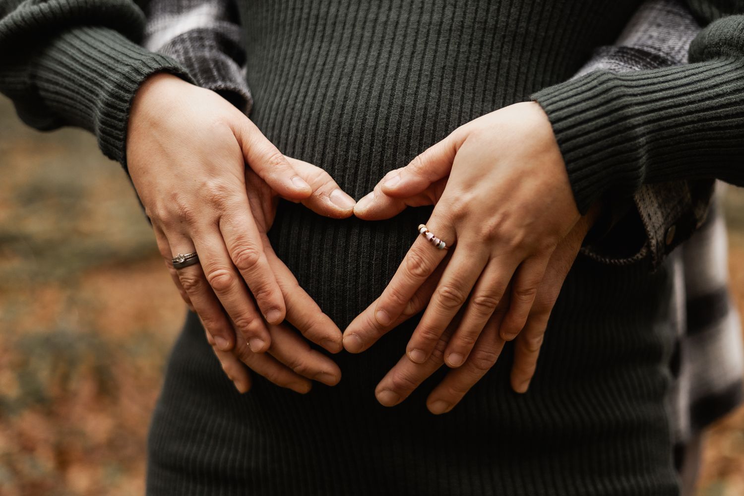 Hände formen ein Herz über dem Babybauch, der einen dunklen Strickpullover trägt.