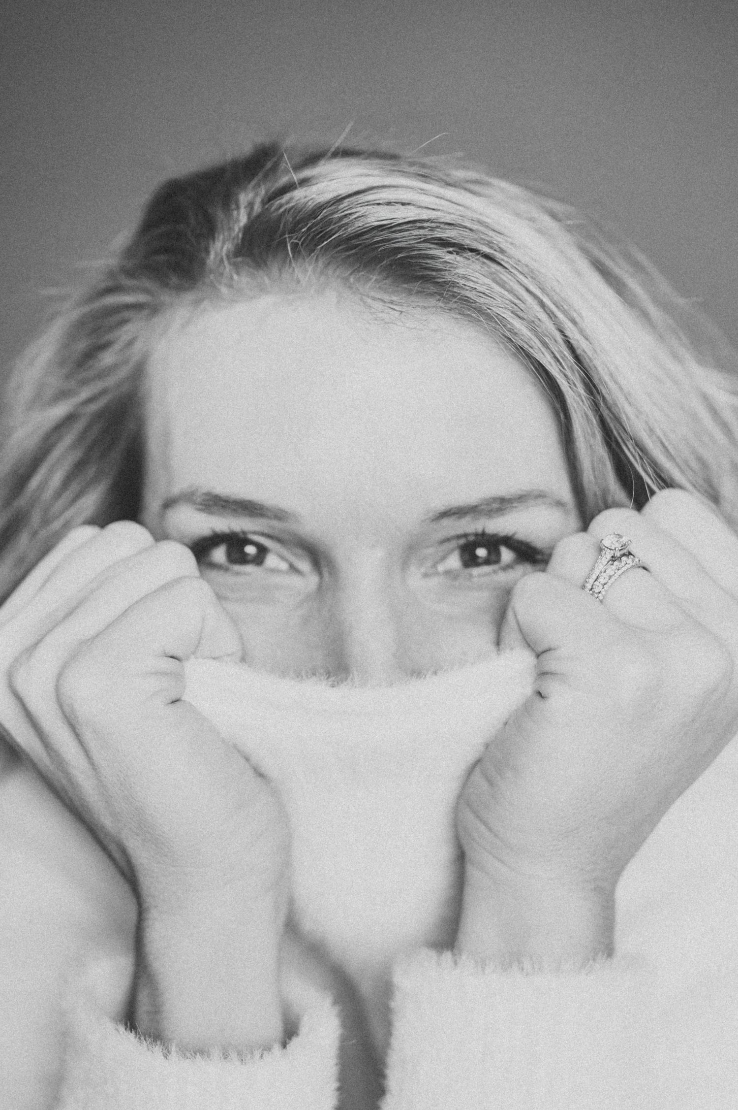 Black and white portrait photograph showing someone's eyes peeking over a cozy white sweater pulled up to cover their nose.
