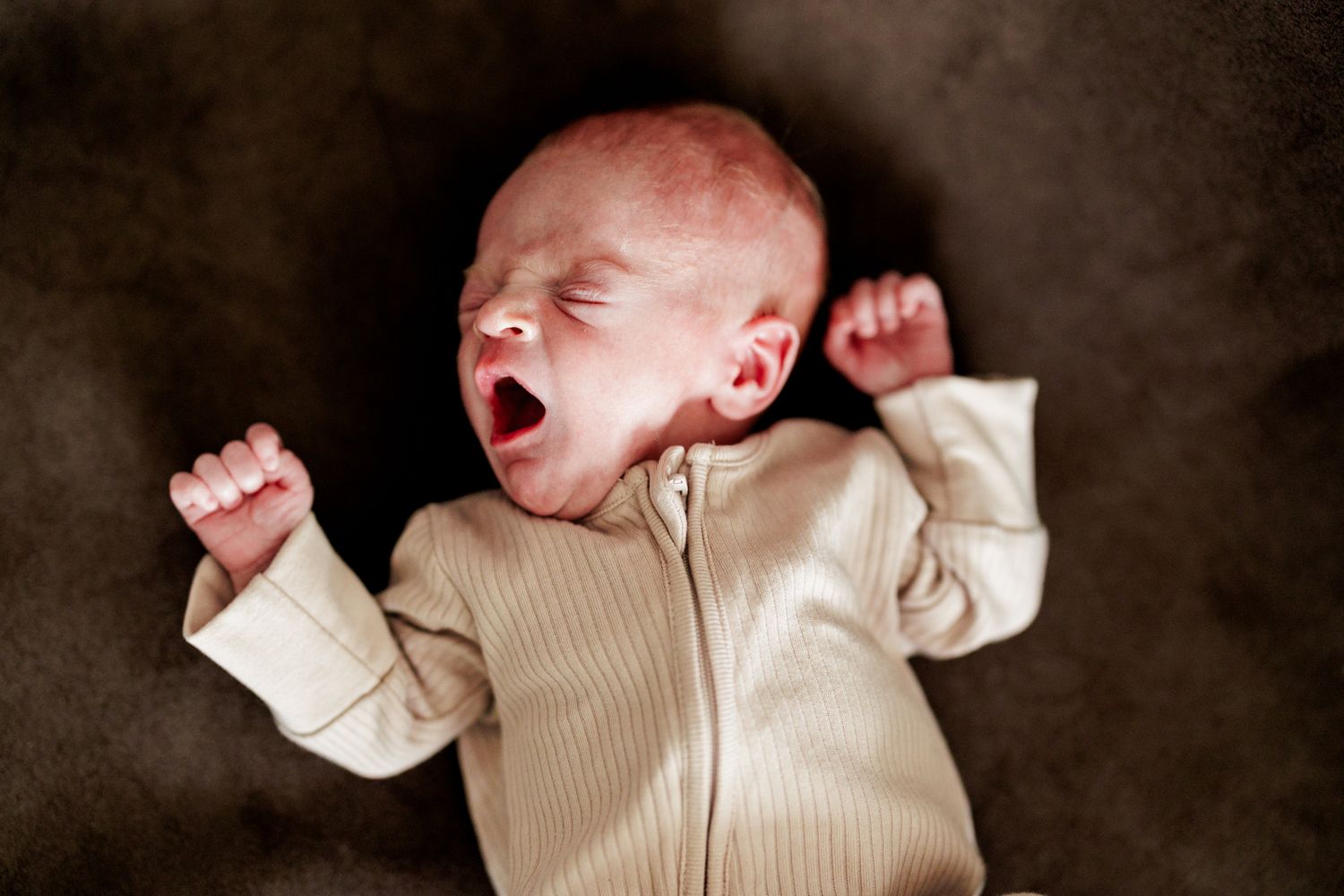 A newborn baby with eyes closed, mouth open, and fists clenched, wearing a beige outfit.