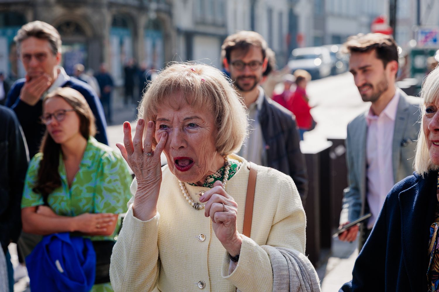 An elderly woman with blonde hair is shown in a crowd of people on a city street, with a surprised expression on her face.