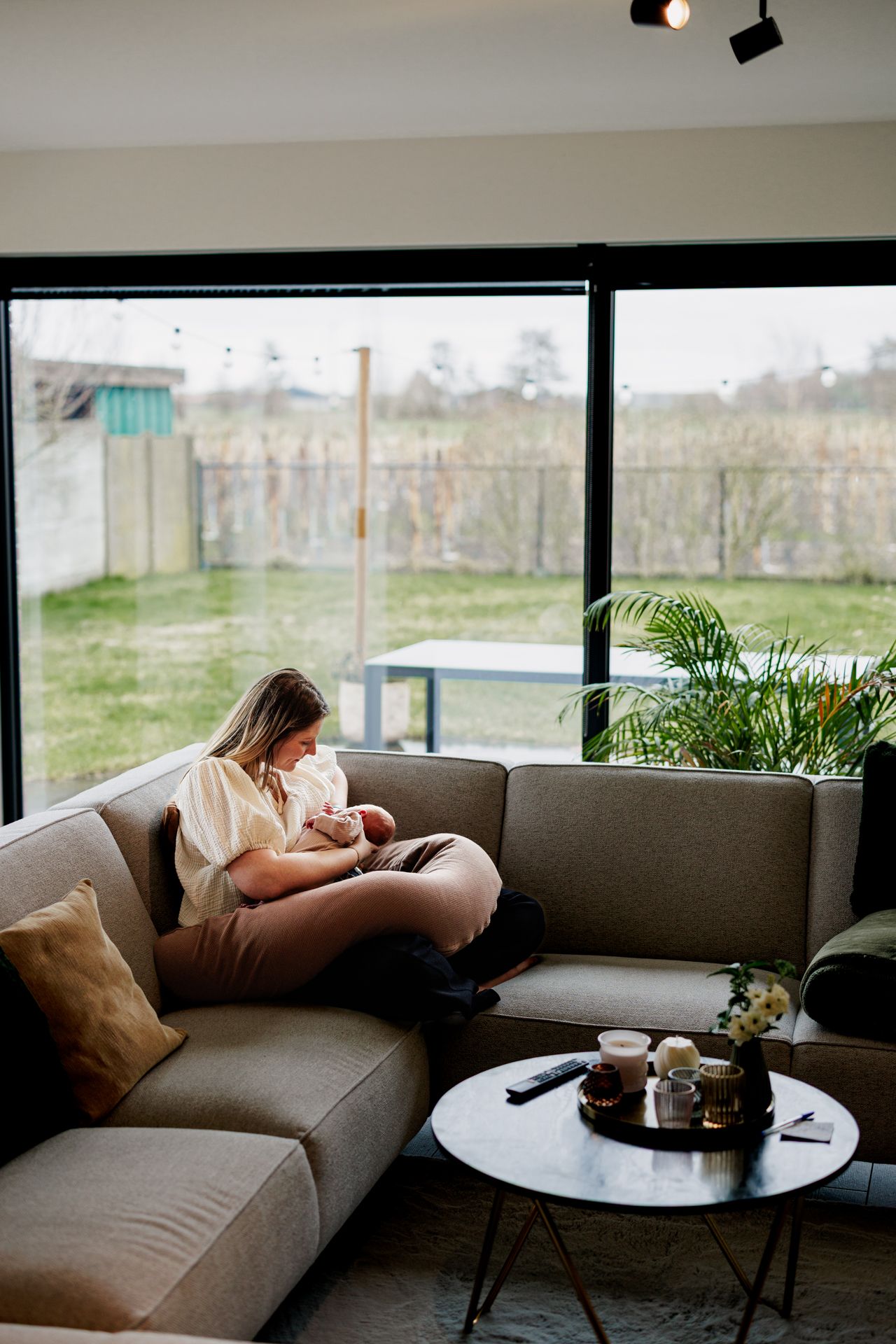 A woman relaxes on a couch in a cozy living room with large windows overlooking a backyard.