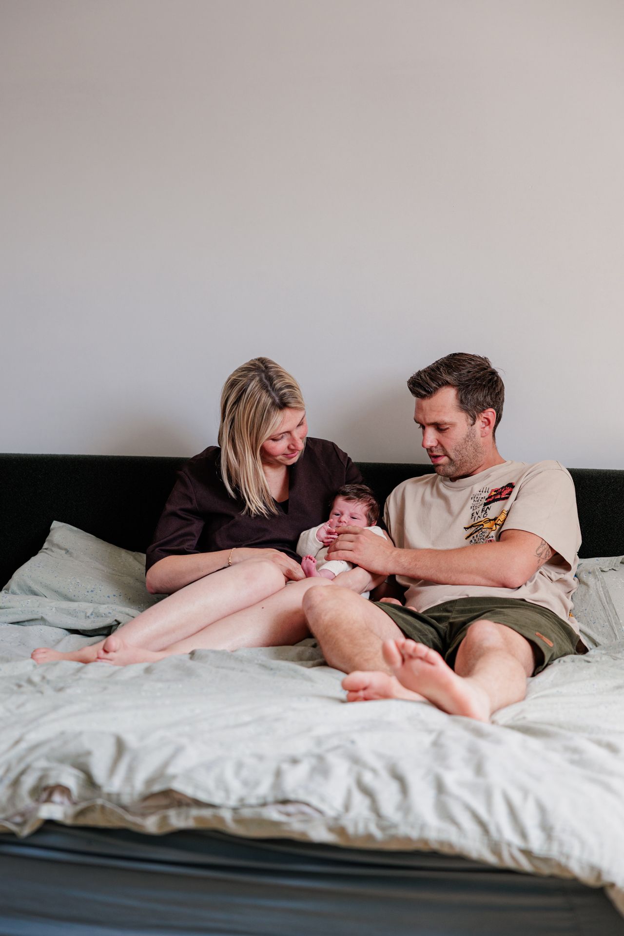 A couple sitting on a bed together, lovingly looking at their newborn baby in a cozy bedroom setting.