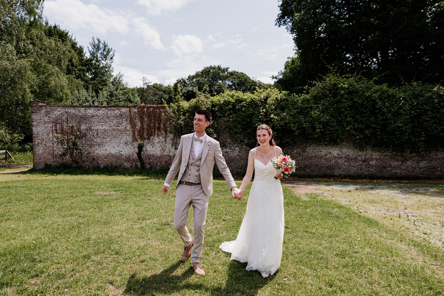 A bride and groom walk together on a grassy lawn surrounded by trees and a stone wall.