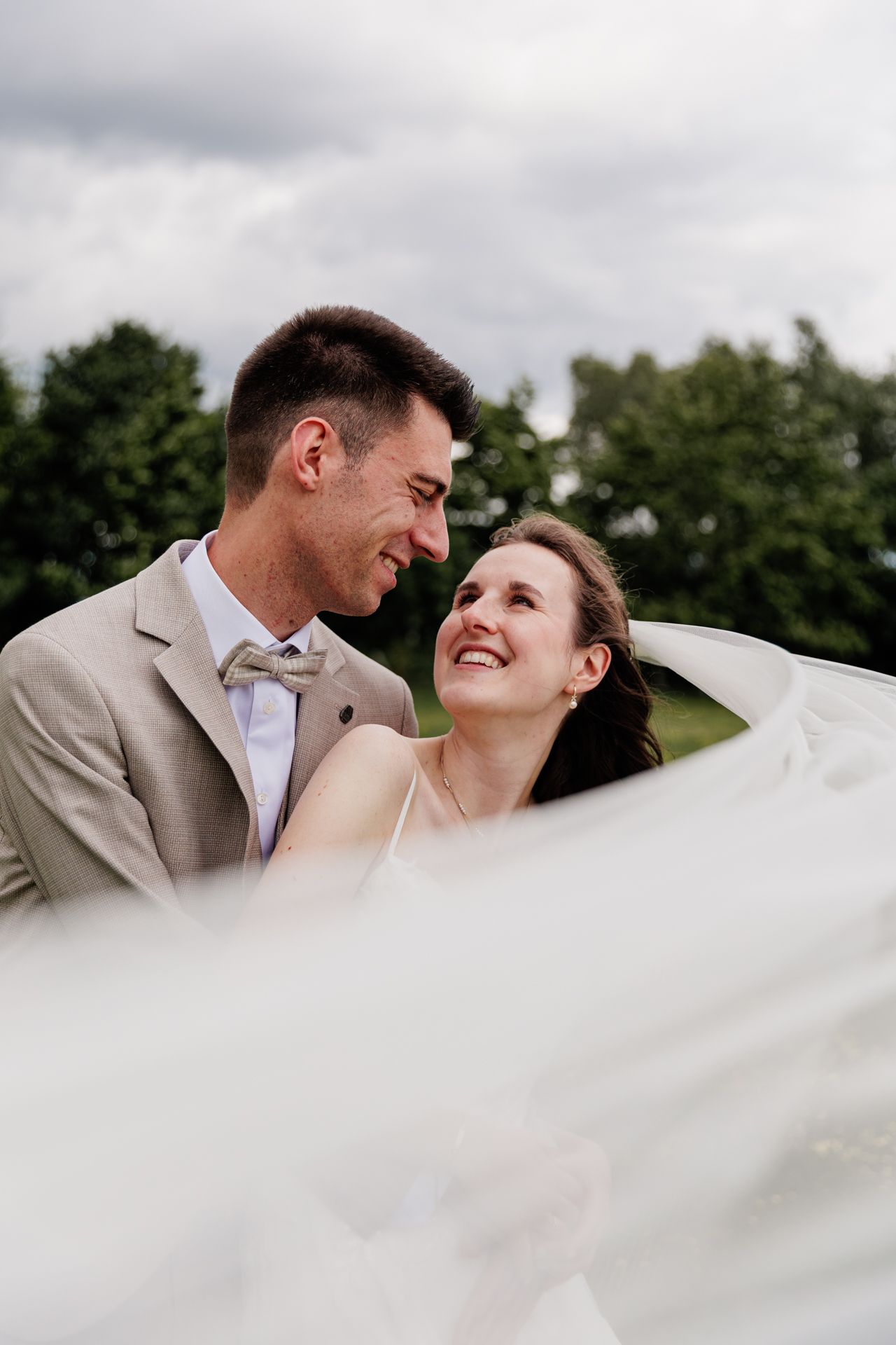 A newlywed couple embraces and smiles at each other in a serene outdoor setting with trees and greenery in the background.