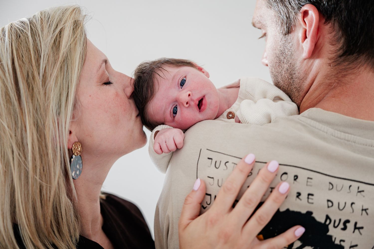 A mother kisses her newborn baby held by the father in a tender family portrait photo.