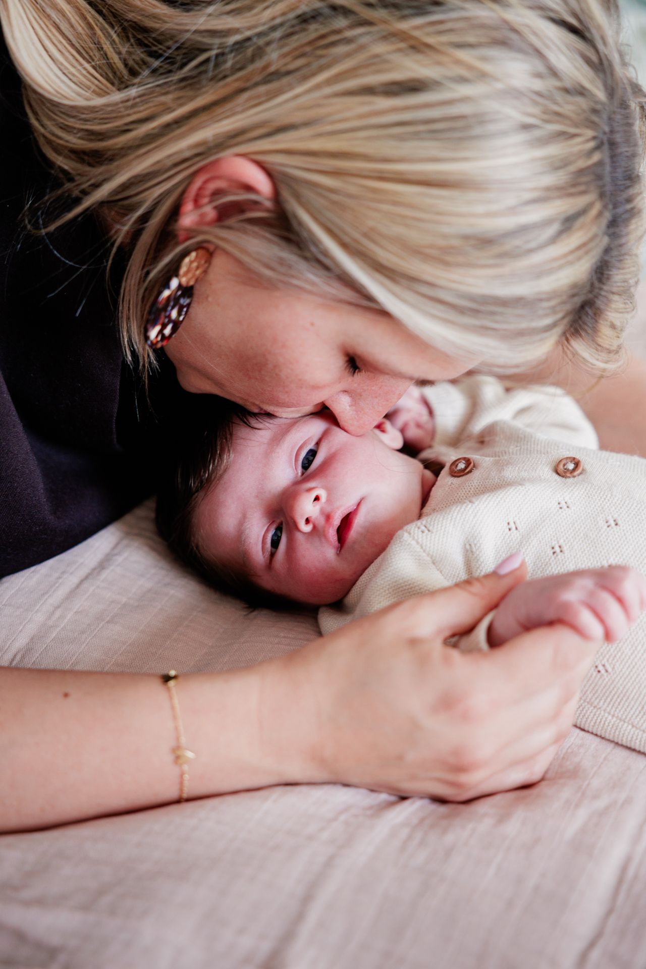 Blonde mother gently kissing her newborn baby wearing a cream knit onesie while lying on a soft pink blanket.