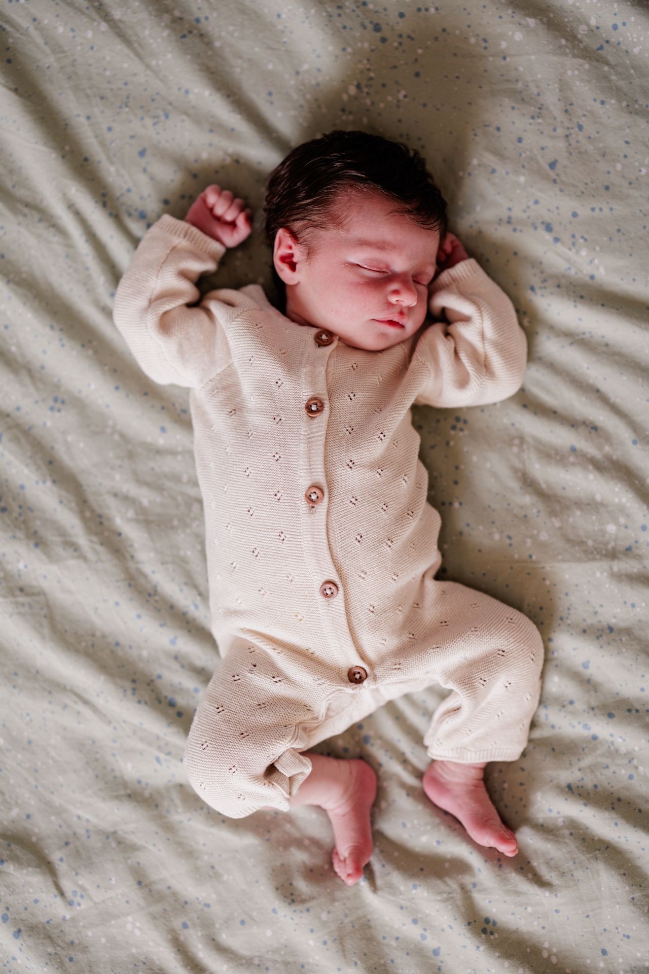 Peaceful newborn baby sleeping on a soft blanket wearing a cream knit button-up onesie with tiny bare feet.