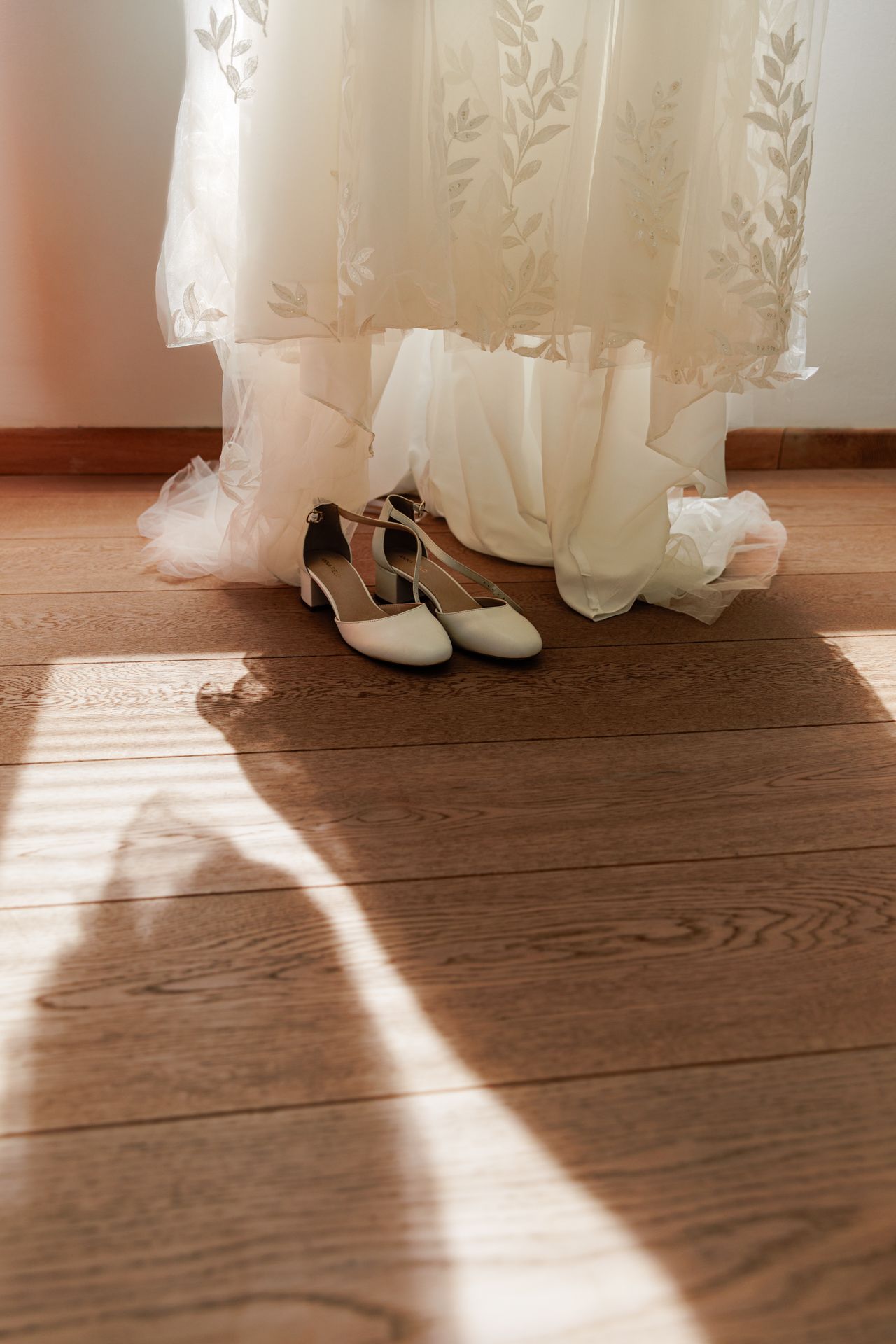 The image shows a pair of white high-heeled shoes on a wooden floor, with a white lace wedding dress visible in the background.