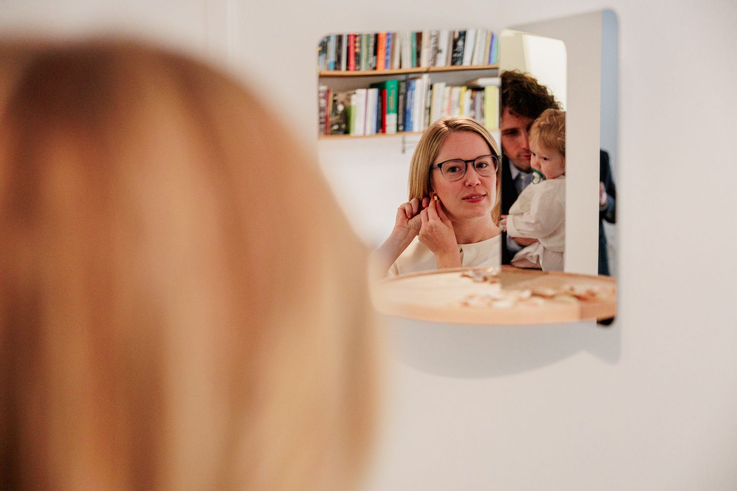 A person with glasses is reflected in a mirror, appearing to be in a home or office environment.