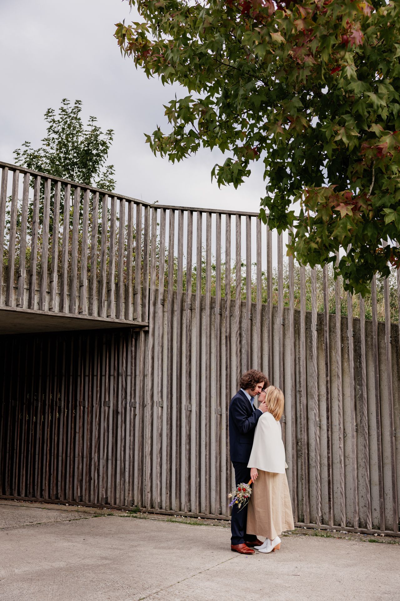A bride and groom embracing in front of a wooden fence with trees in the background.