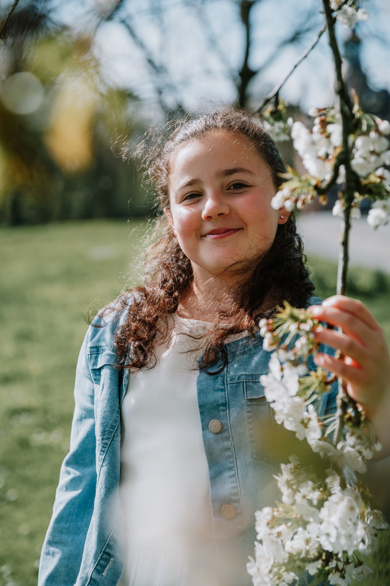 A young girl with curly hair stands among blooming flowers, smiling brightly.