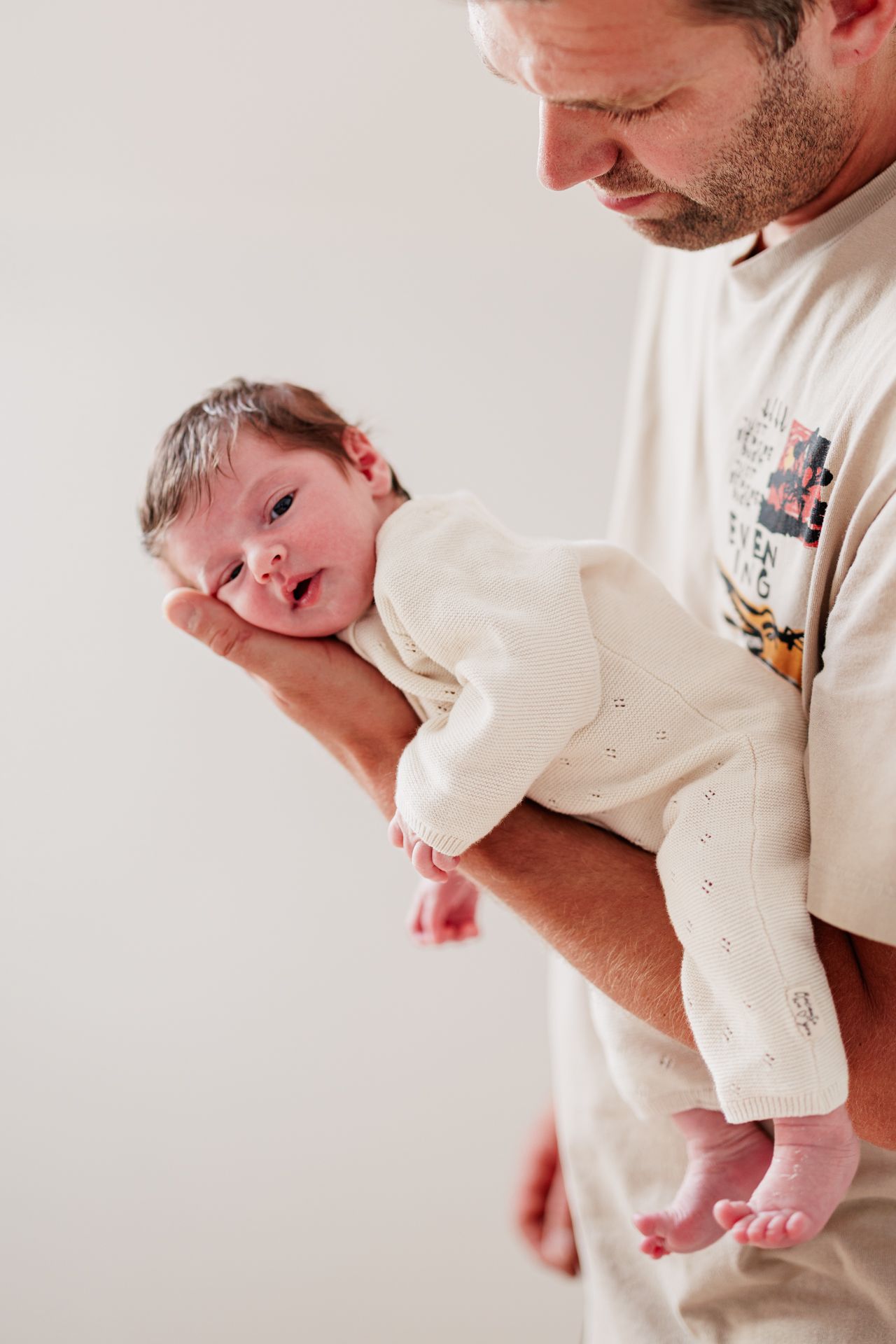 A bearded father tenderly holds his newborn baby in a cream outfit against a white background.