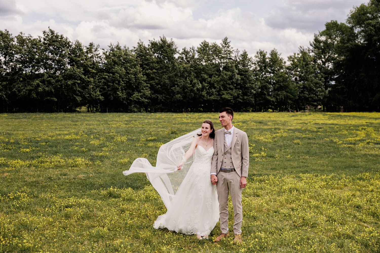 A bride and groom stand in a grassy field surrounded by trees on their wedding day.