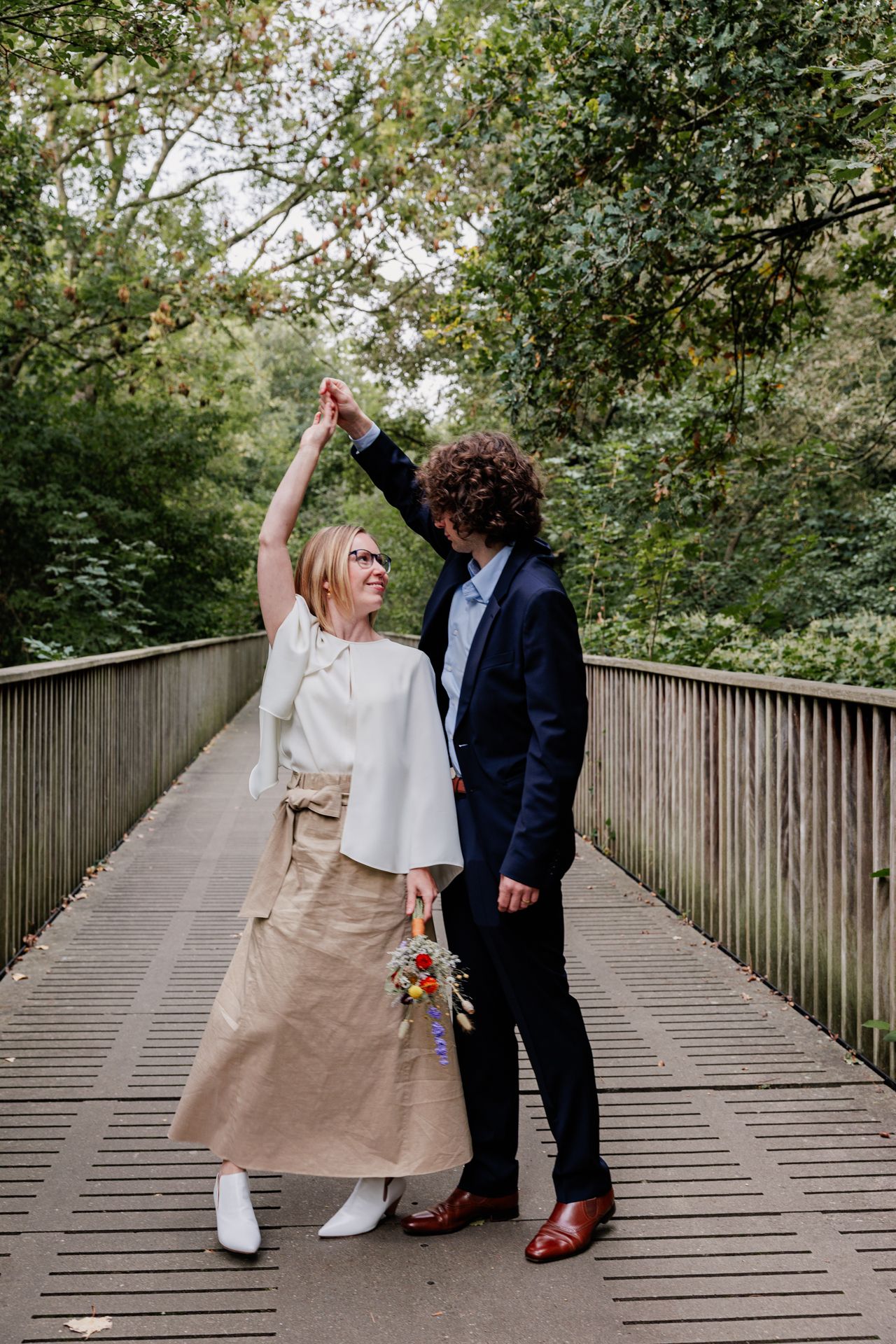 A bride and groom stand on a wooden bridge surrounded by lush greenery, celebrating their wedding day.
