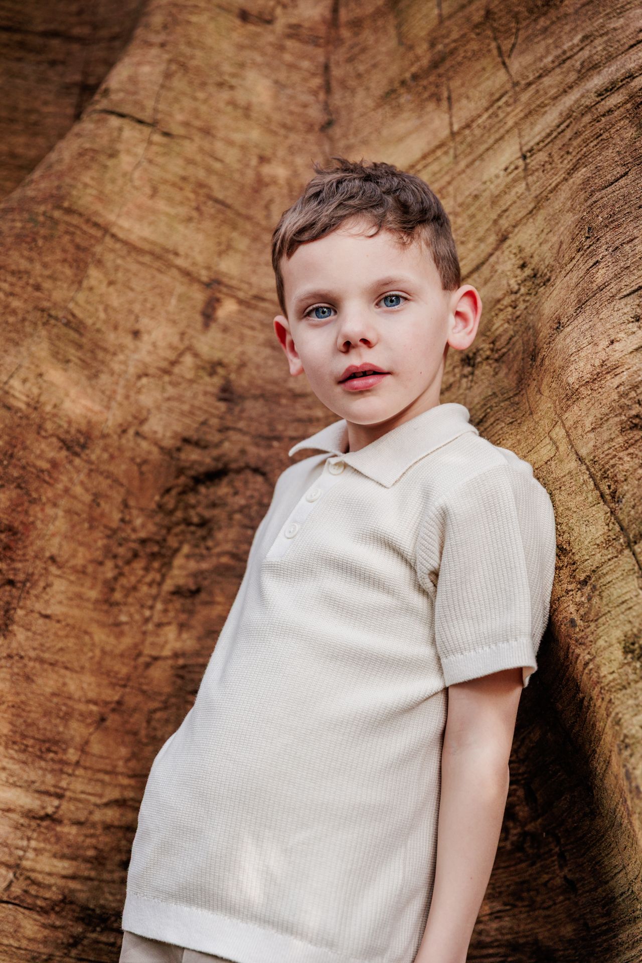 A young boy wearing a white shirt stands in front of a wooden background.