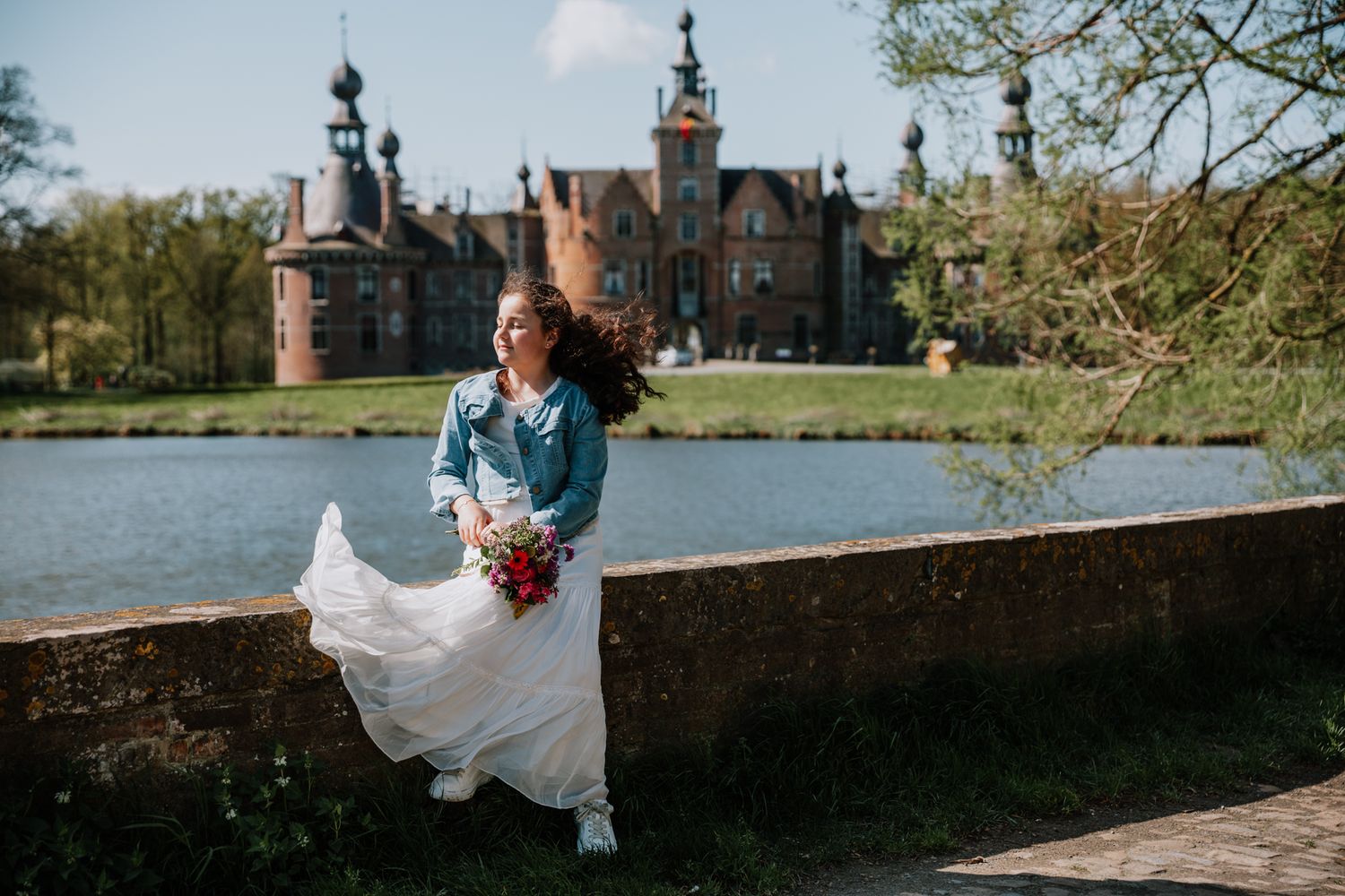 A woman in a white wedding dress stands in front of a historic castle with a pond in the foreground.