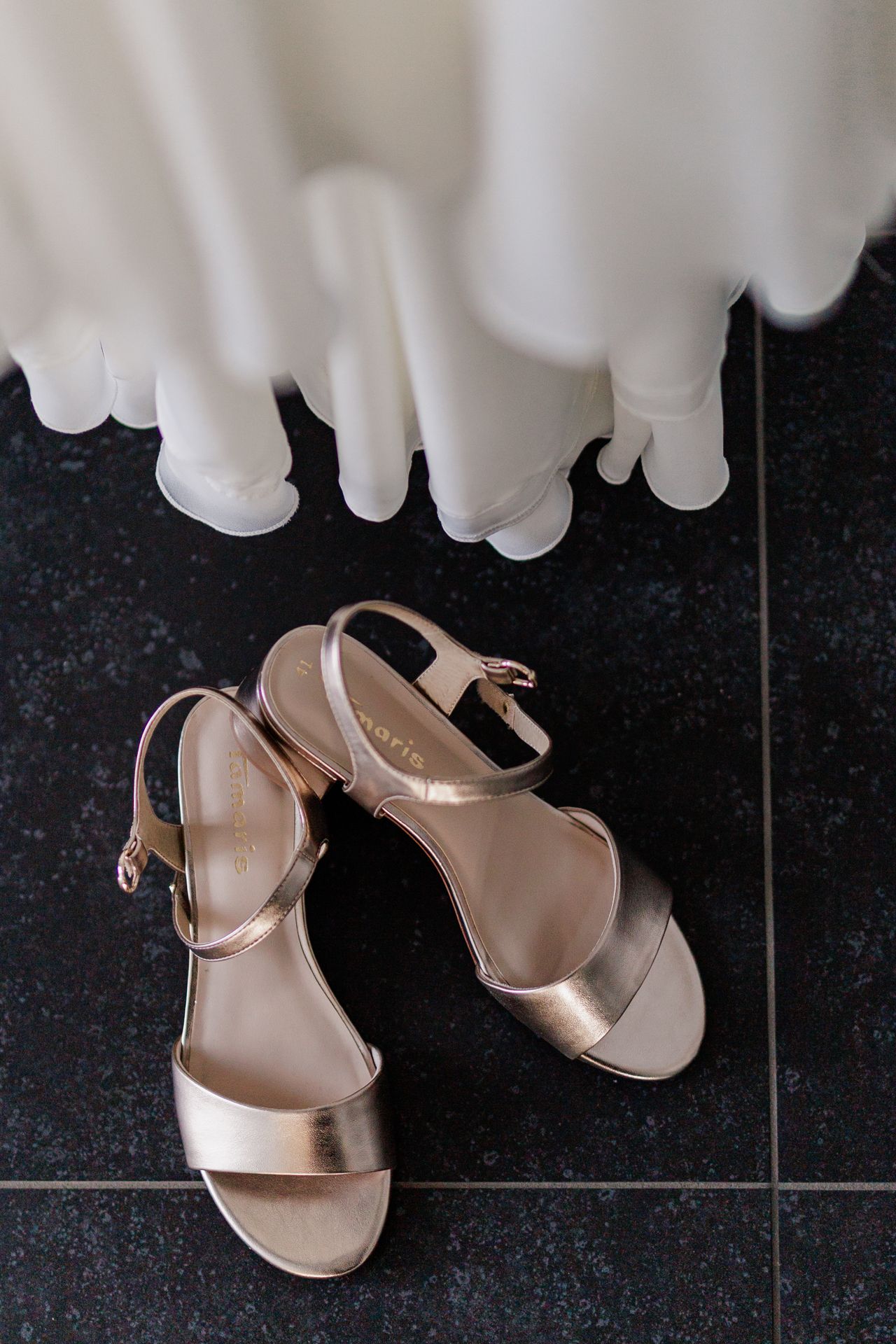A pair of beige high-heel sandals with straps are shown on a black tile floor, with a white dress in the background.