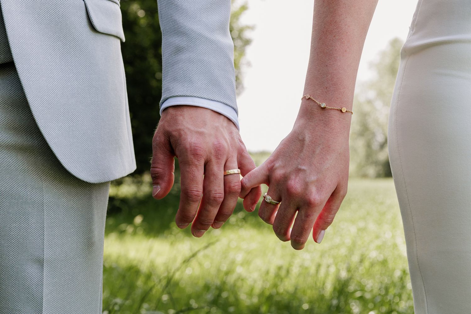 Two hands of a couple holding each other in a grassy outdoor setting.