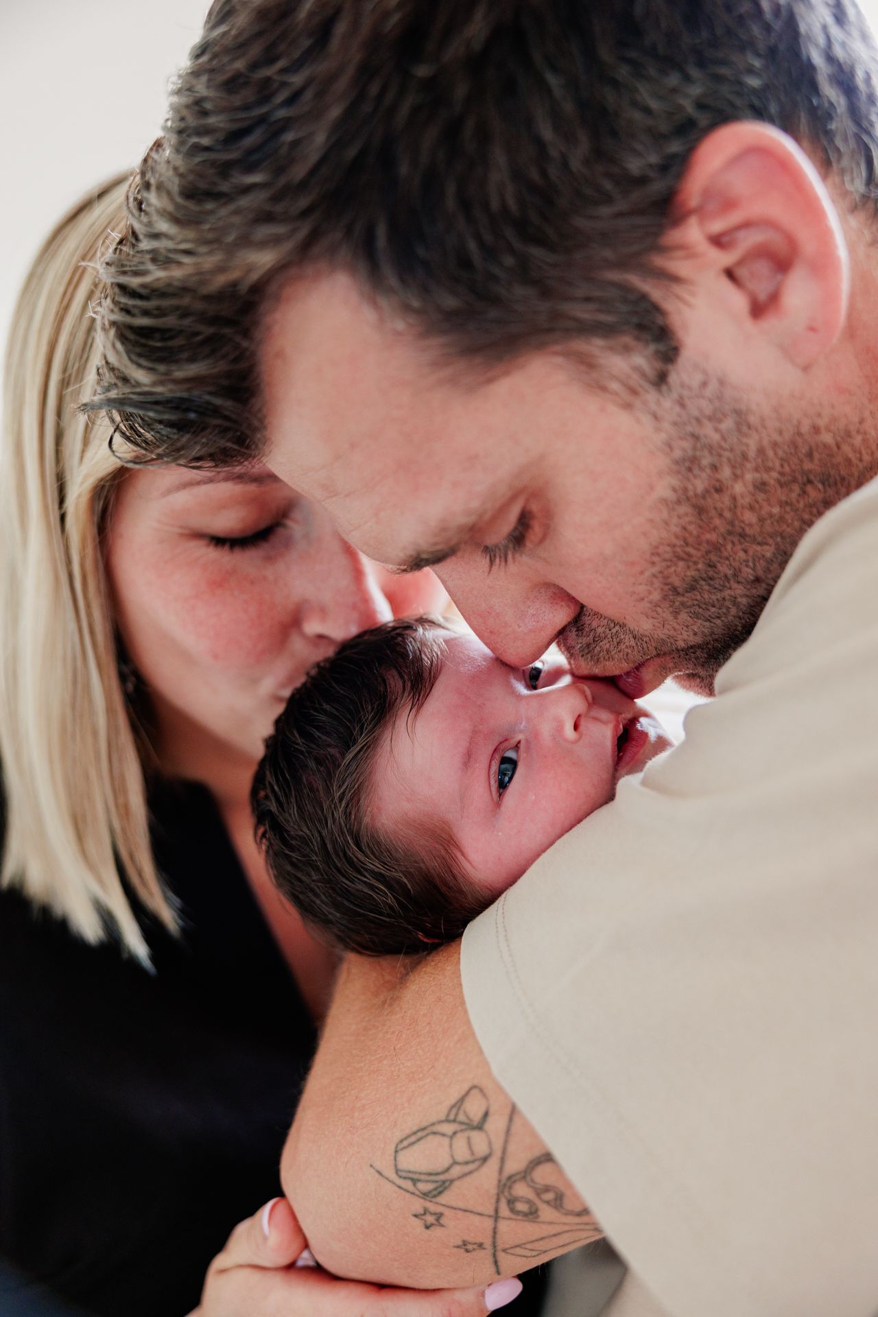 Two loving parents kissing their newborn baby on the forehead, father showing a tattoo on his arm.