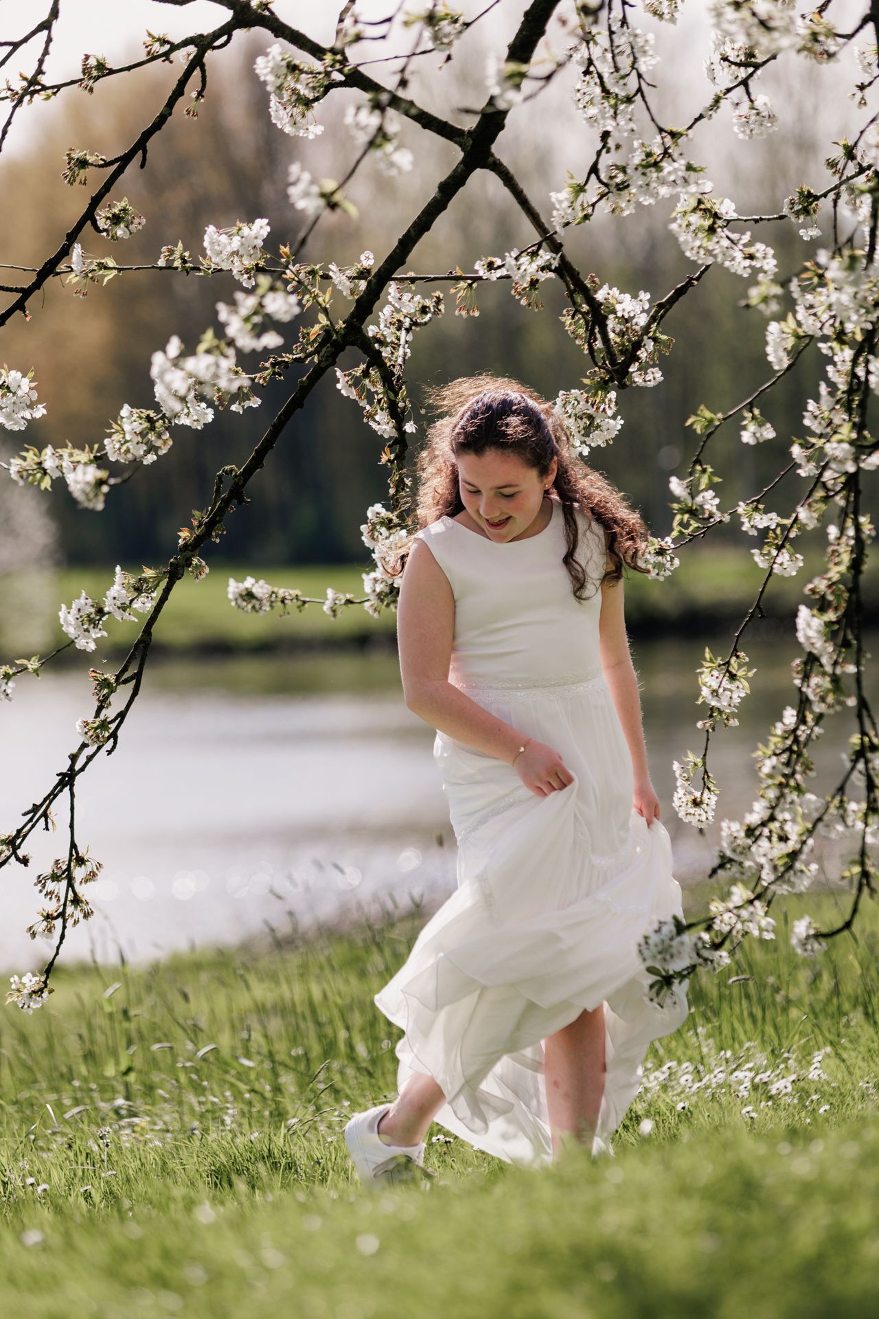 A young woman in a white dress stands among blooming cherry blossom trees in a grassy field.