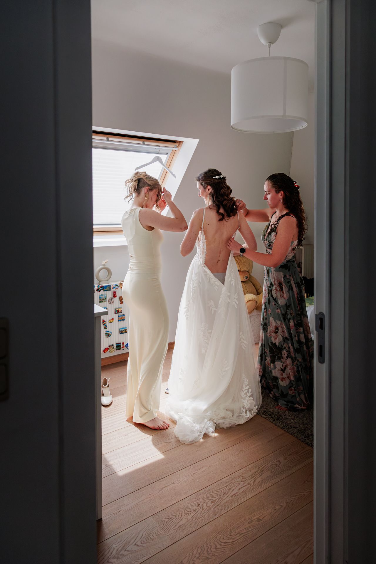 A bride getting ready for her wedding, assisted by two bridesmaids in a room with natural light.