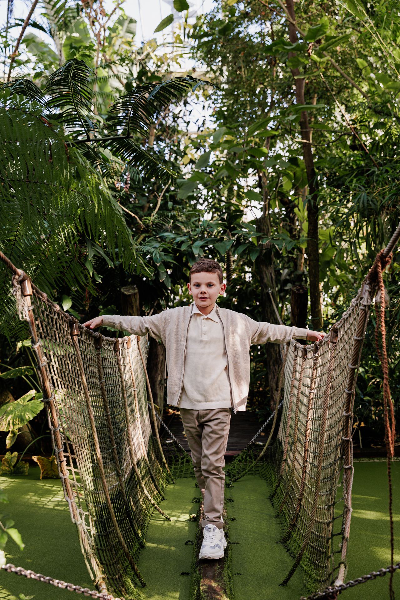 A young man stands on a rope bridge in a lush, tropical garden with palm trees and greenery surrounding him.