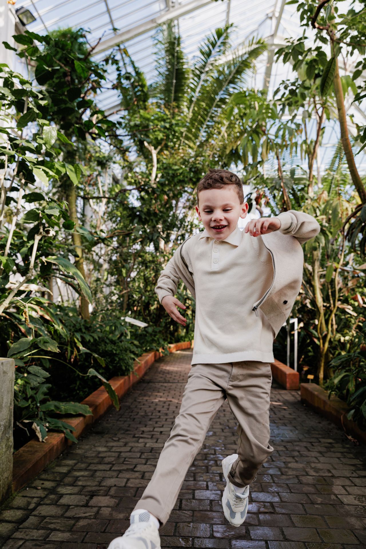 A young boy is walking on a brick path in a greenhouse filled with lush, tropical plants.