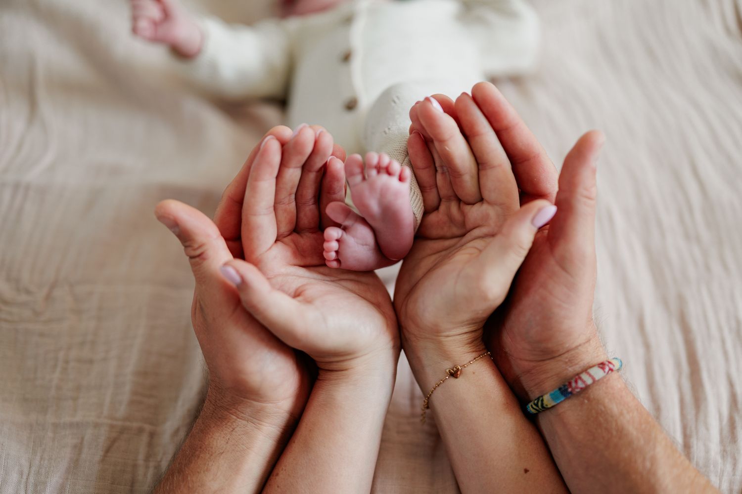 Two parents cup their hands together holding tiny newborn baby feet on a soft white blanket.