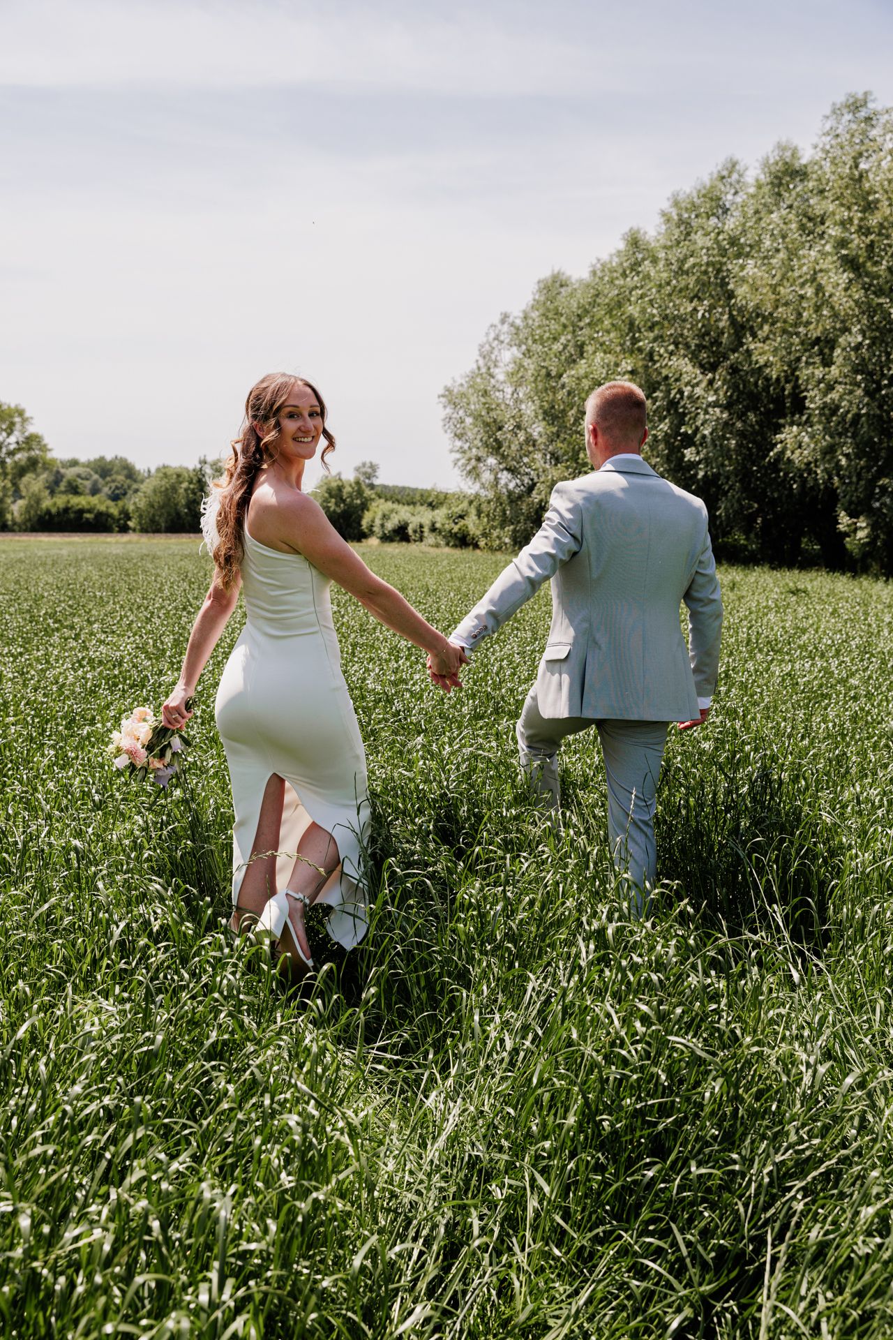 A bride and groom walk hand-in-hand through a lush green field on their wedding day.