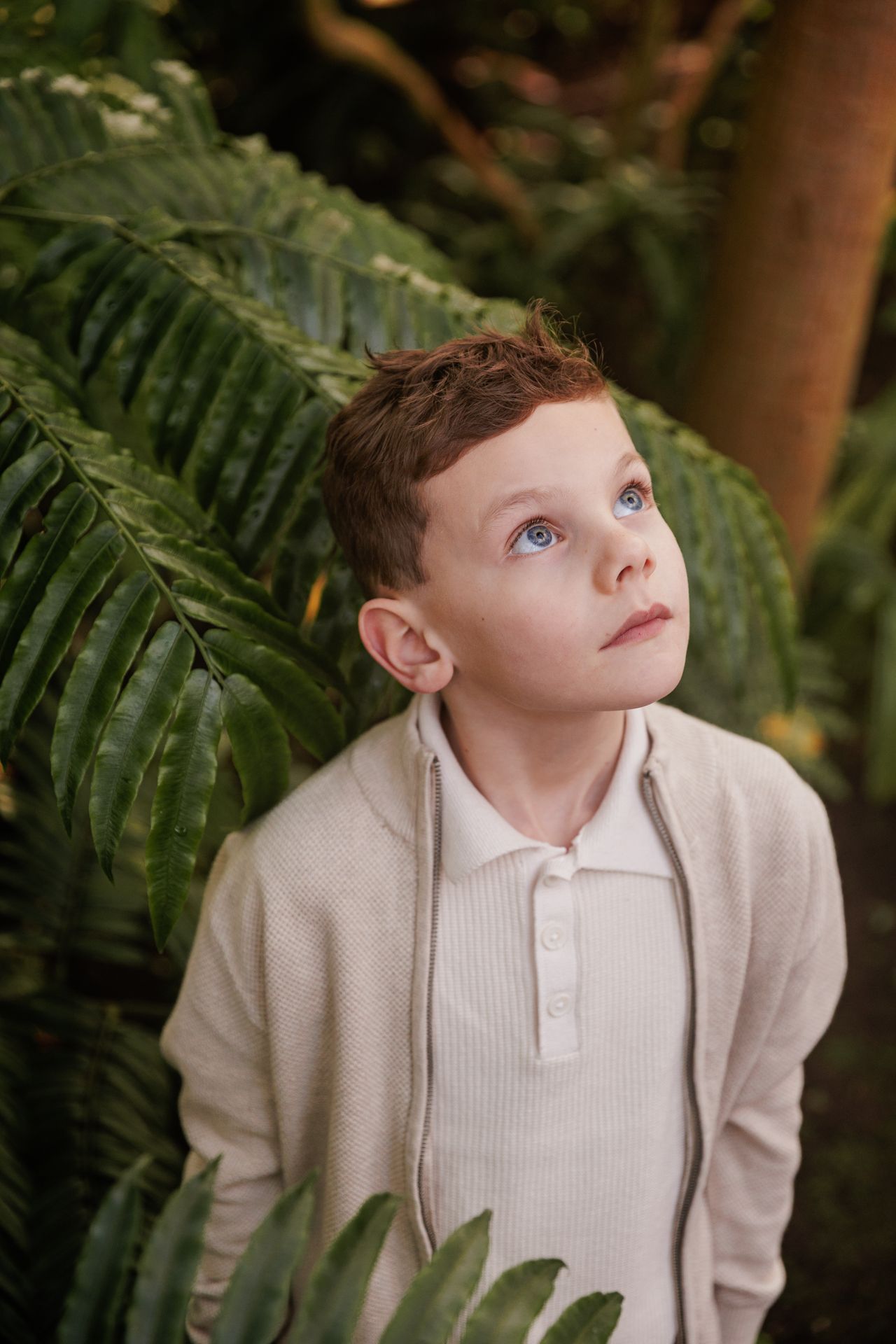 A young boy with reddish-brown hair stands among lush green foliage, looking thoughtfully into the distance.
