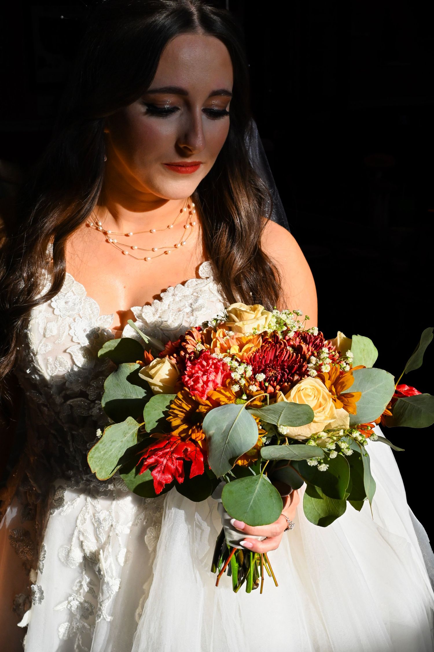A bride in a white lace dress holds a vibrant autumn bouquet with red and yellow flowers against a dark background.