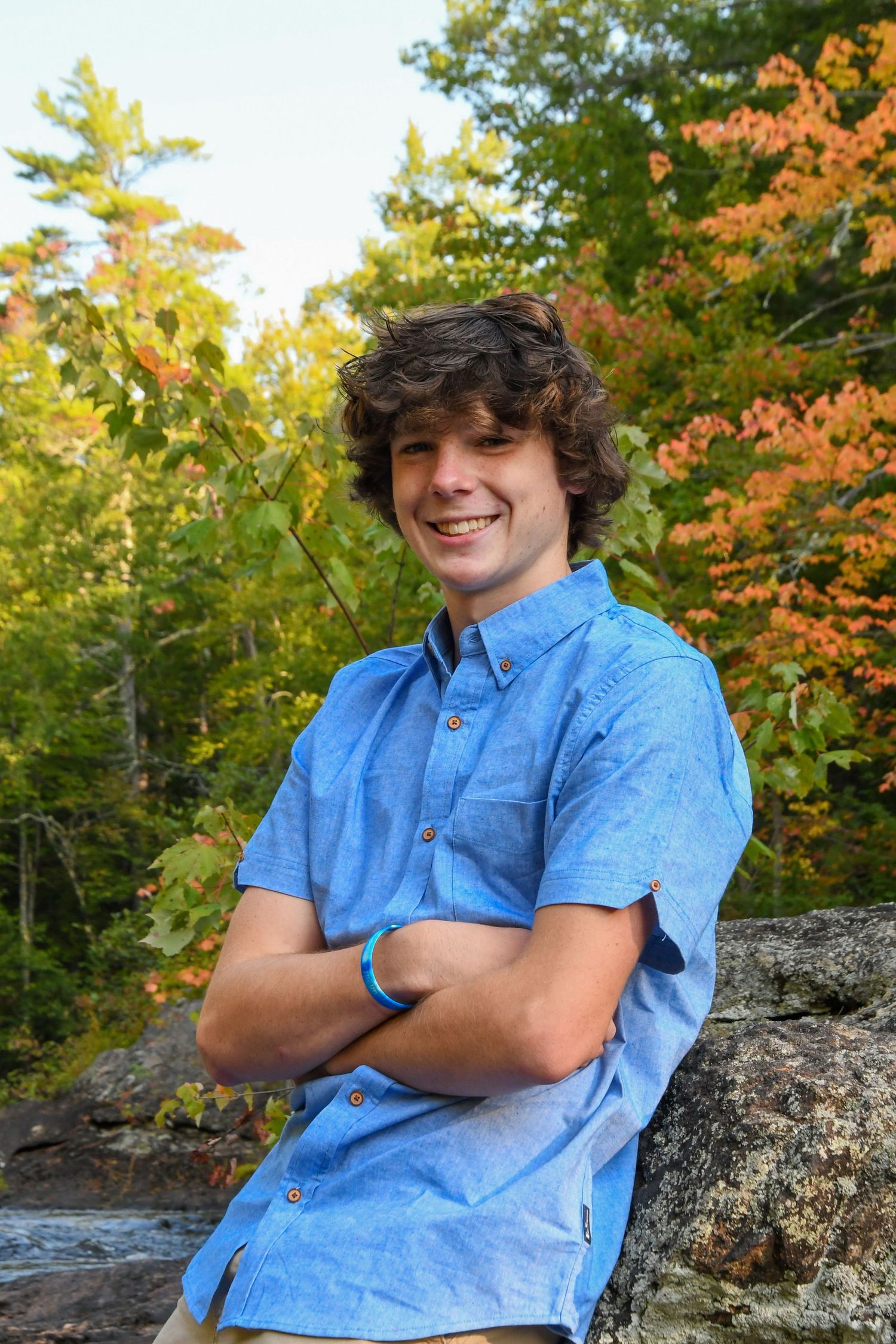 Person in blue button-down shirt poses with crossed arms against autumn foliage background.