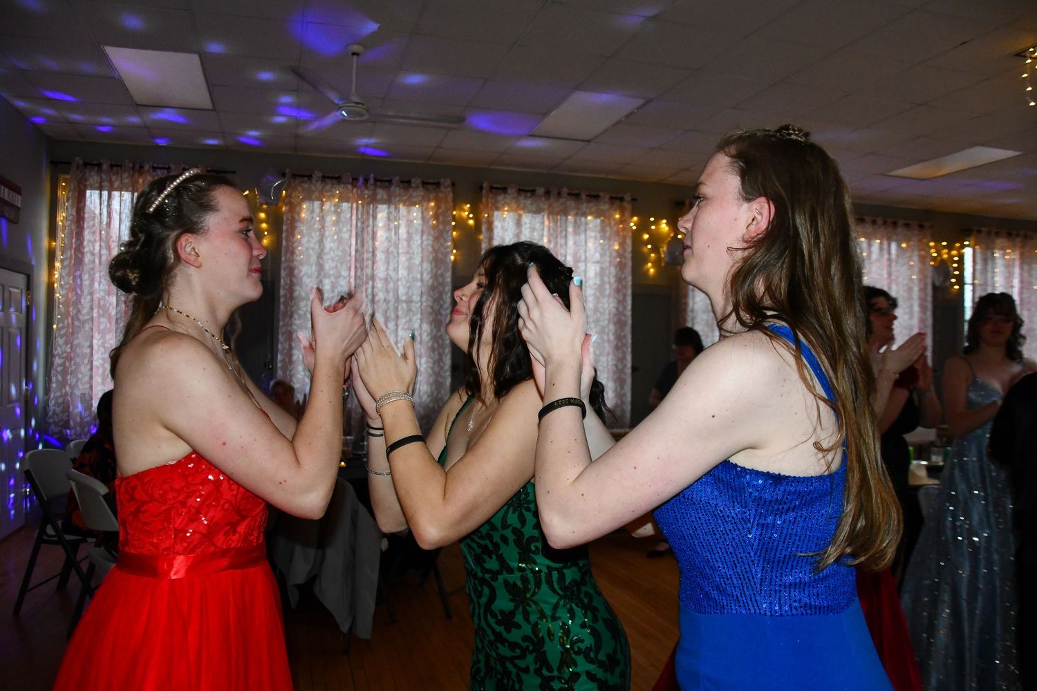 Friends in formal dresses dance together at celebration with blue lighting and decorative backdrop.