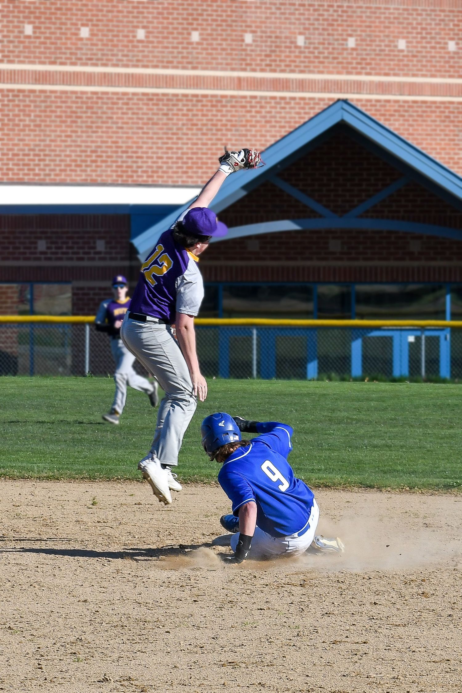 Baseball player leaps to catch a high throw as runner slides into base during game.