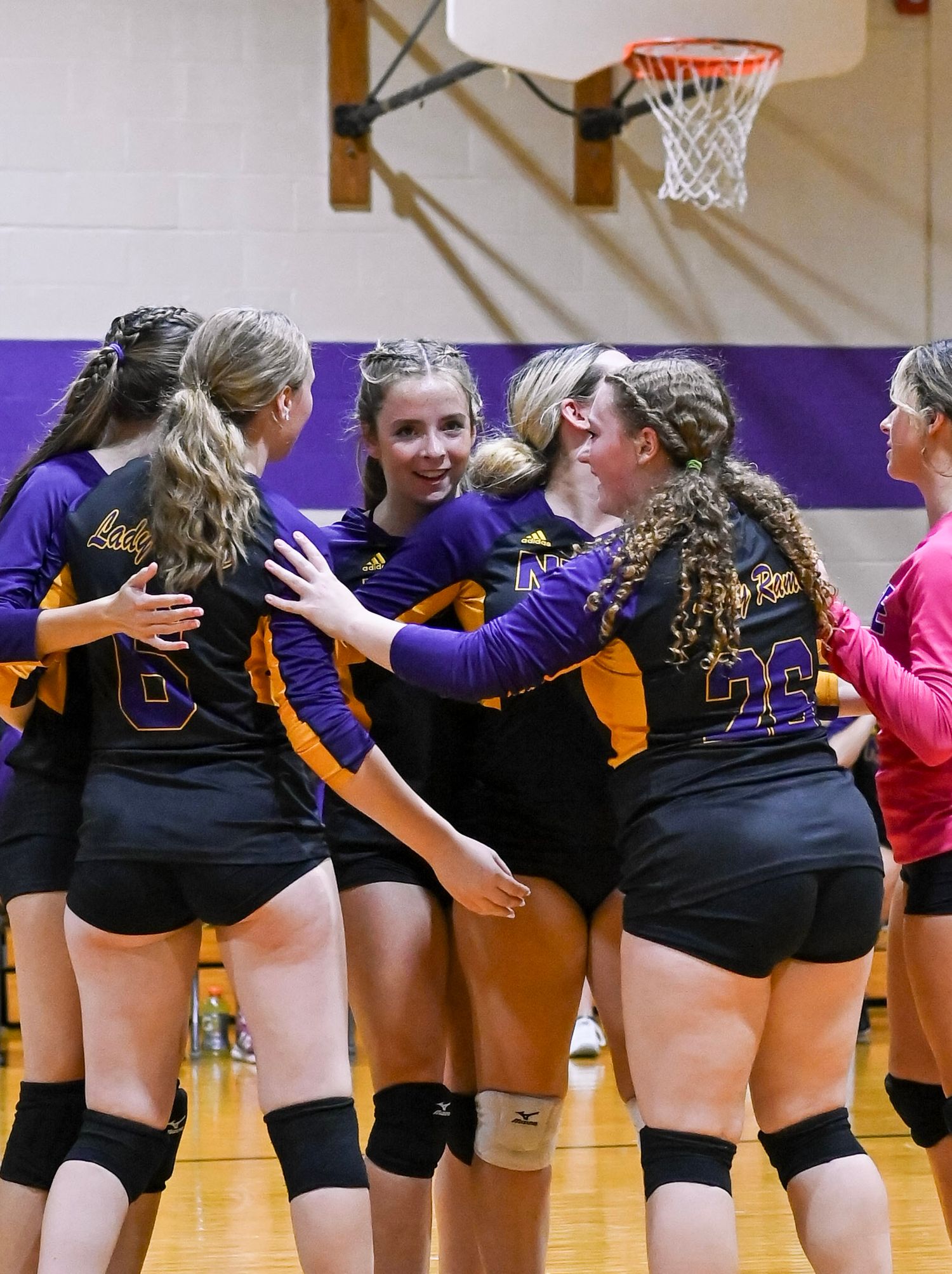 Female volleyball players in black uniforms huddle together in a line on the court during a game celebration.