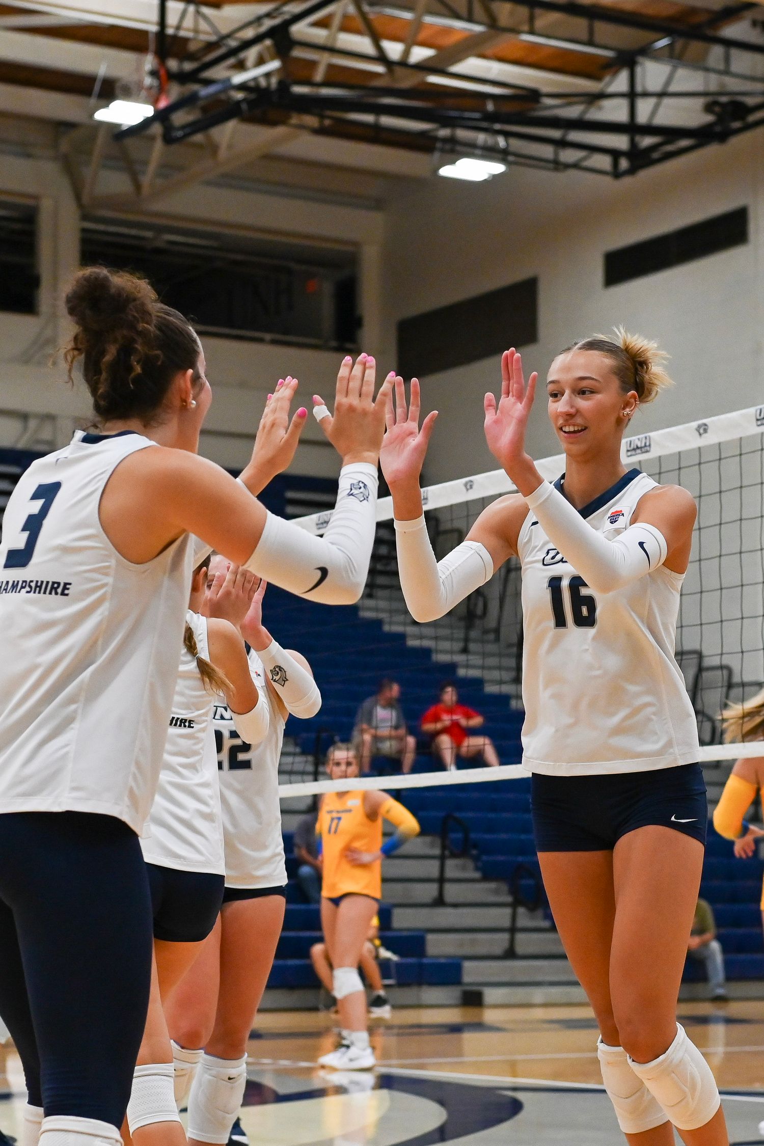 Female volleyball players in white uniforms high-five each other during a game in an indoor gymnasium.