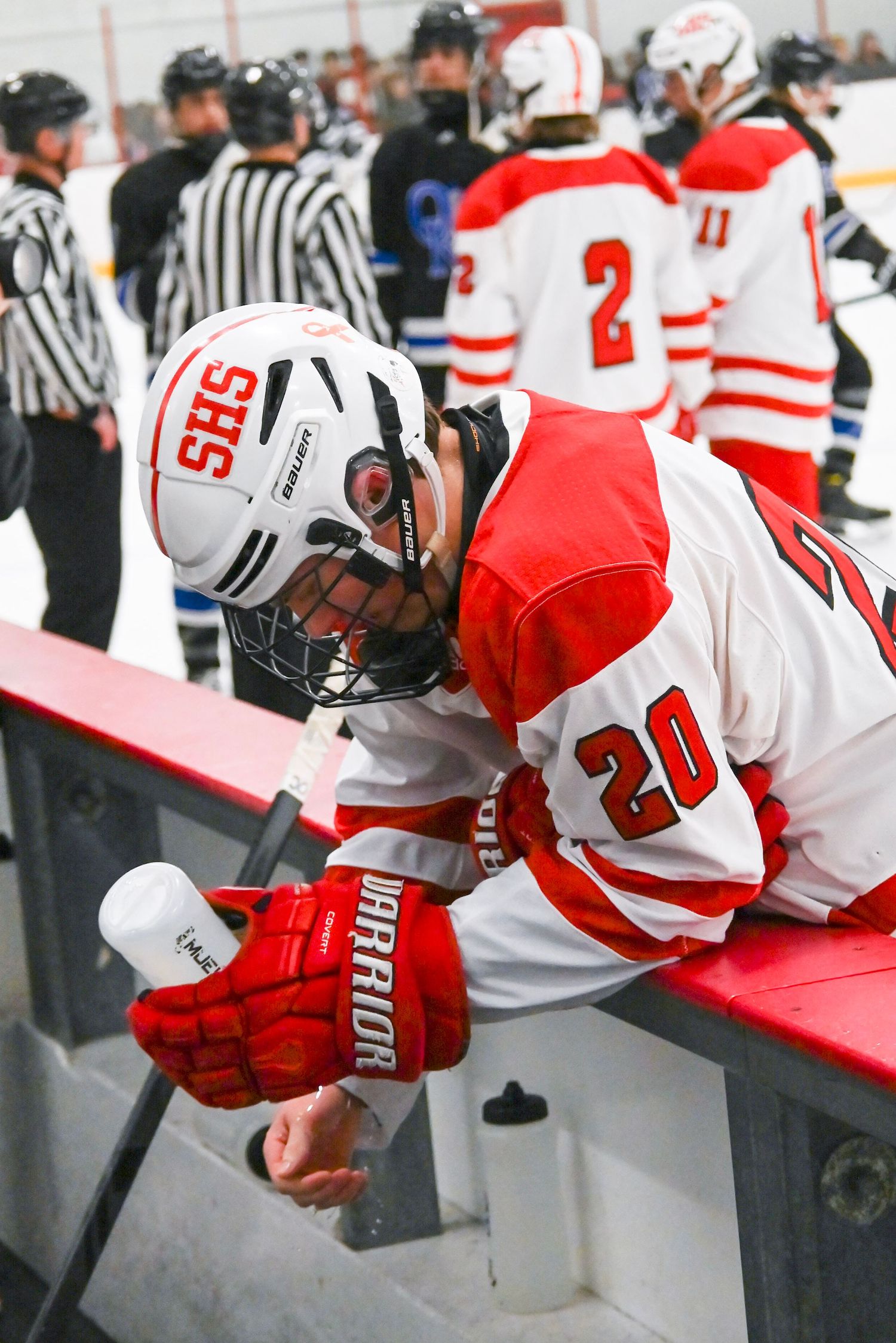 Hockey player in white and red jersey #20 leans over boards while officials and teammates gather in background.
