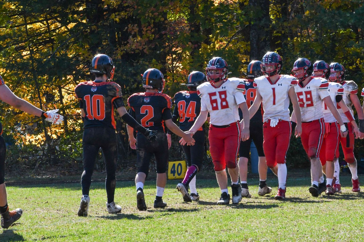 Two football teams line up on field during autumn game with colorful fall foliage in background.