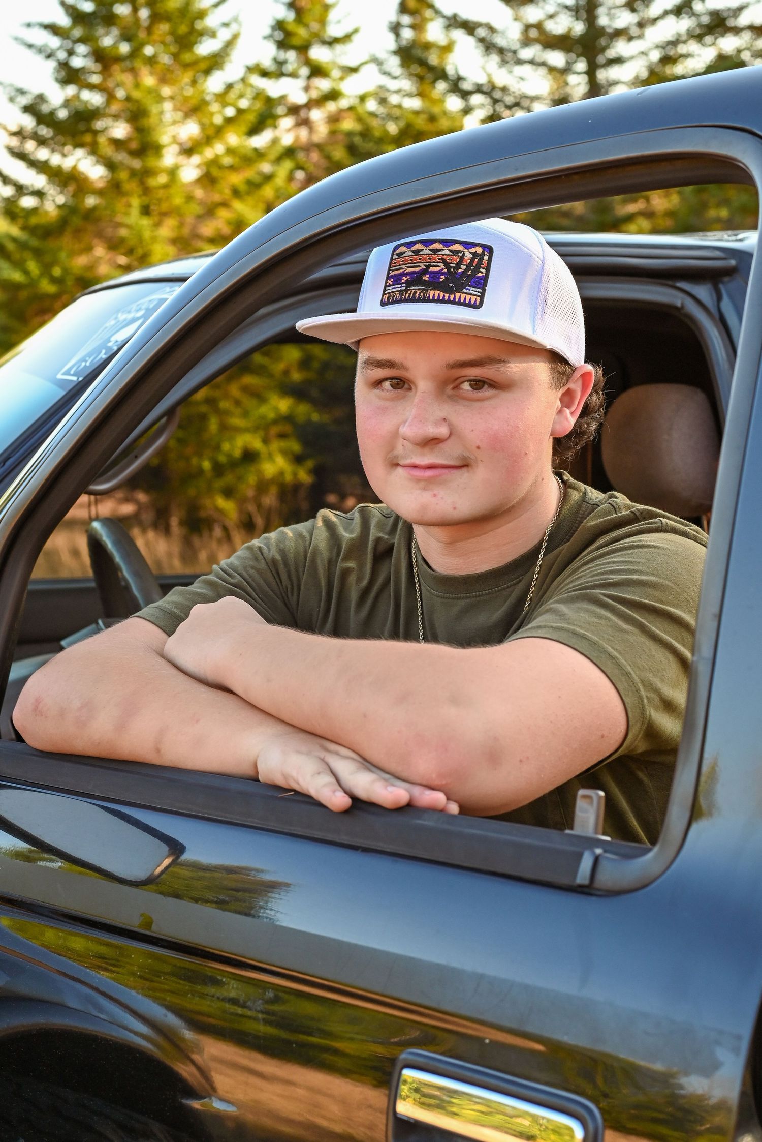 A person sits in a car window wearing a baseball cap and olive green shirt against an evergreen forest background.