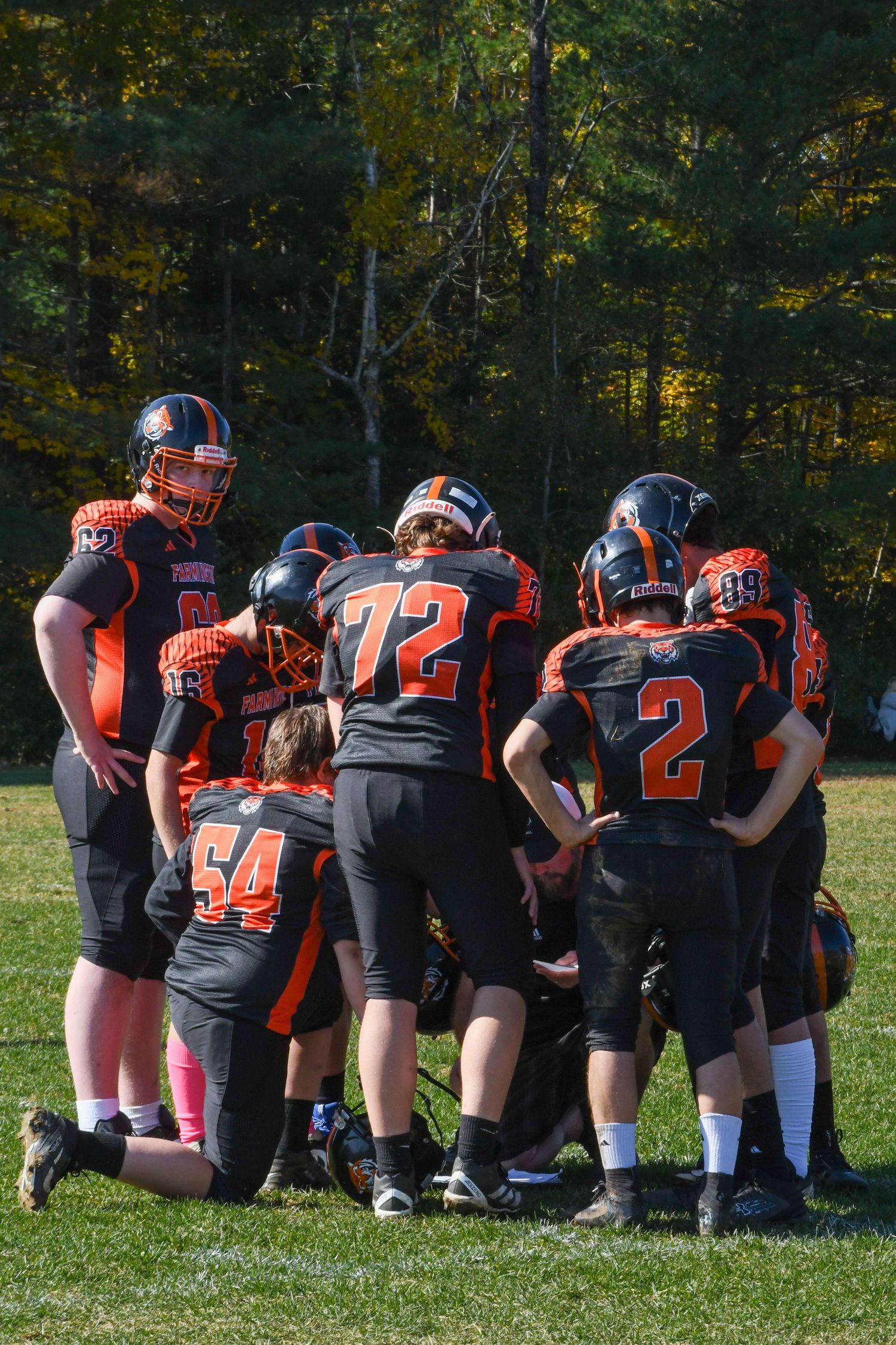 Football players in orange and black uniforms huddle together on a grass field with autumn forest in background.
