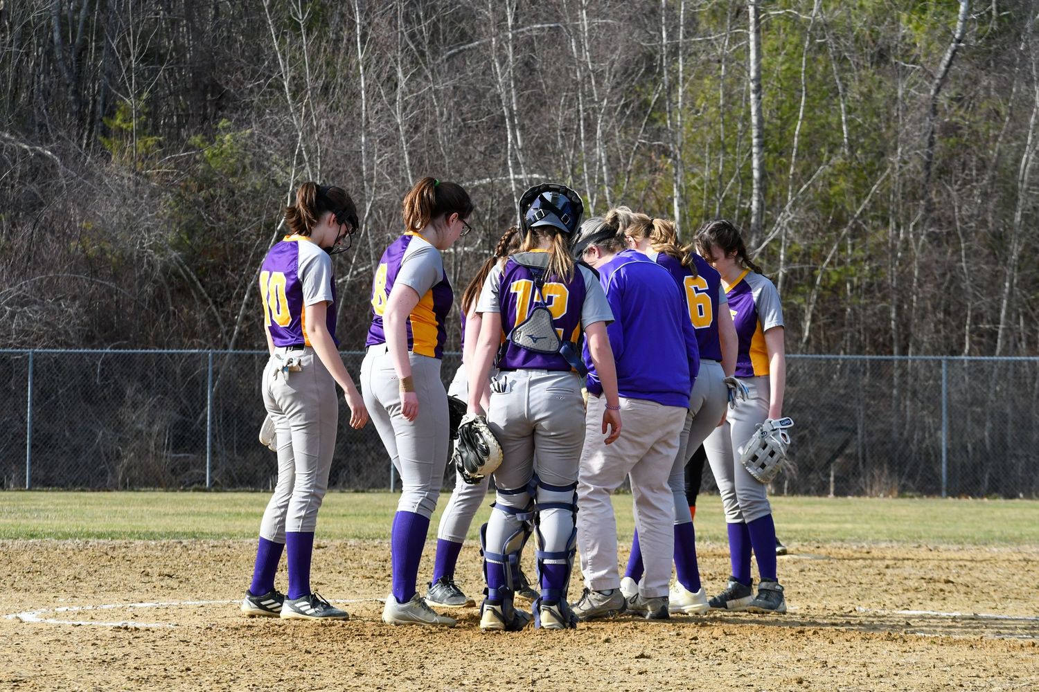 Baseball team huddles together on dirt infield during game while wearing purple and white uniforms.