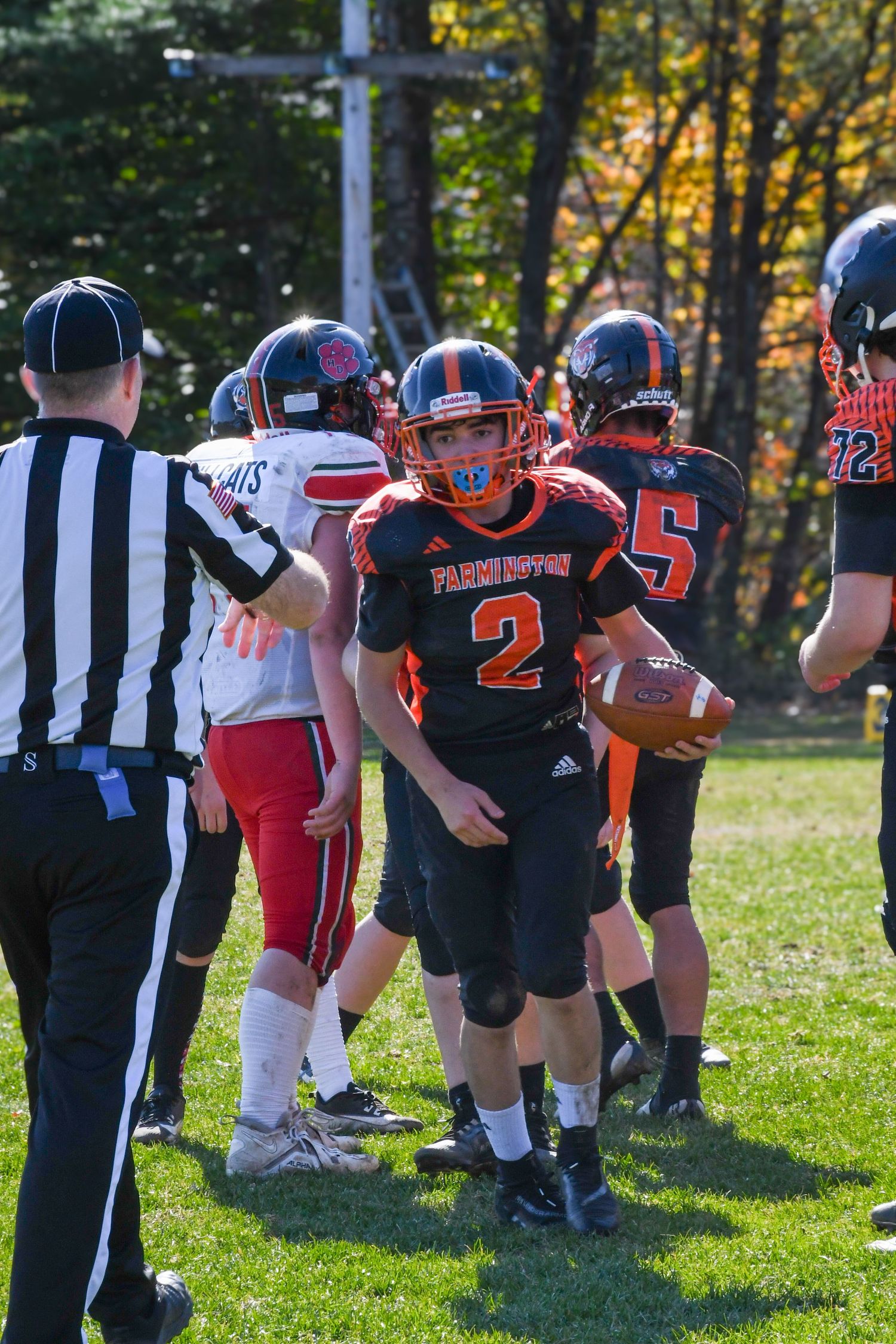 High school football players in orange and black uniforms gather around a referee on a sunny autumn field during a game.