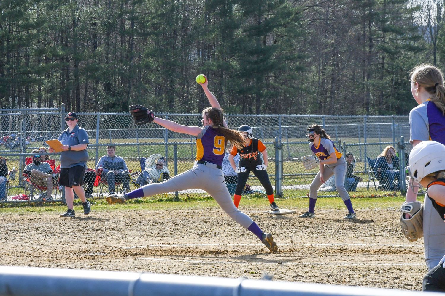Softball pitcher in yellow jersey winds up for powerful pitch on dirt mound during game with spectators watching.