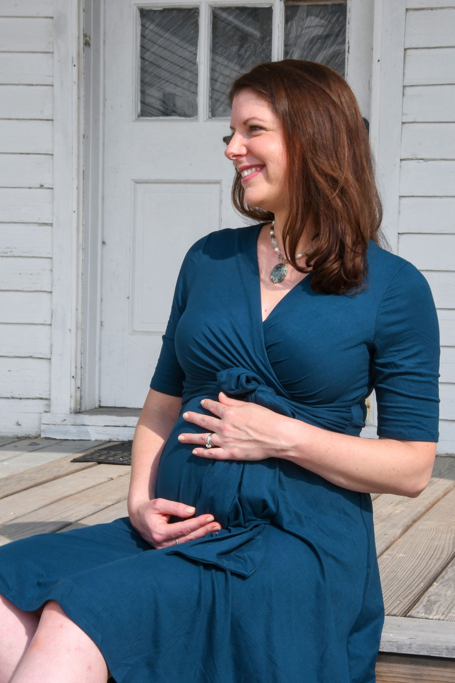 A person wearing a teal blue wrap dress sits on a porch step and smiles while gently touching their stomach.