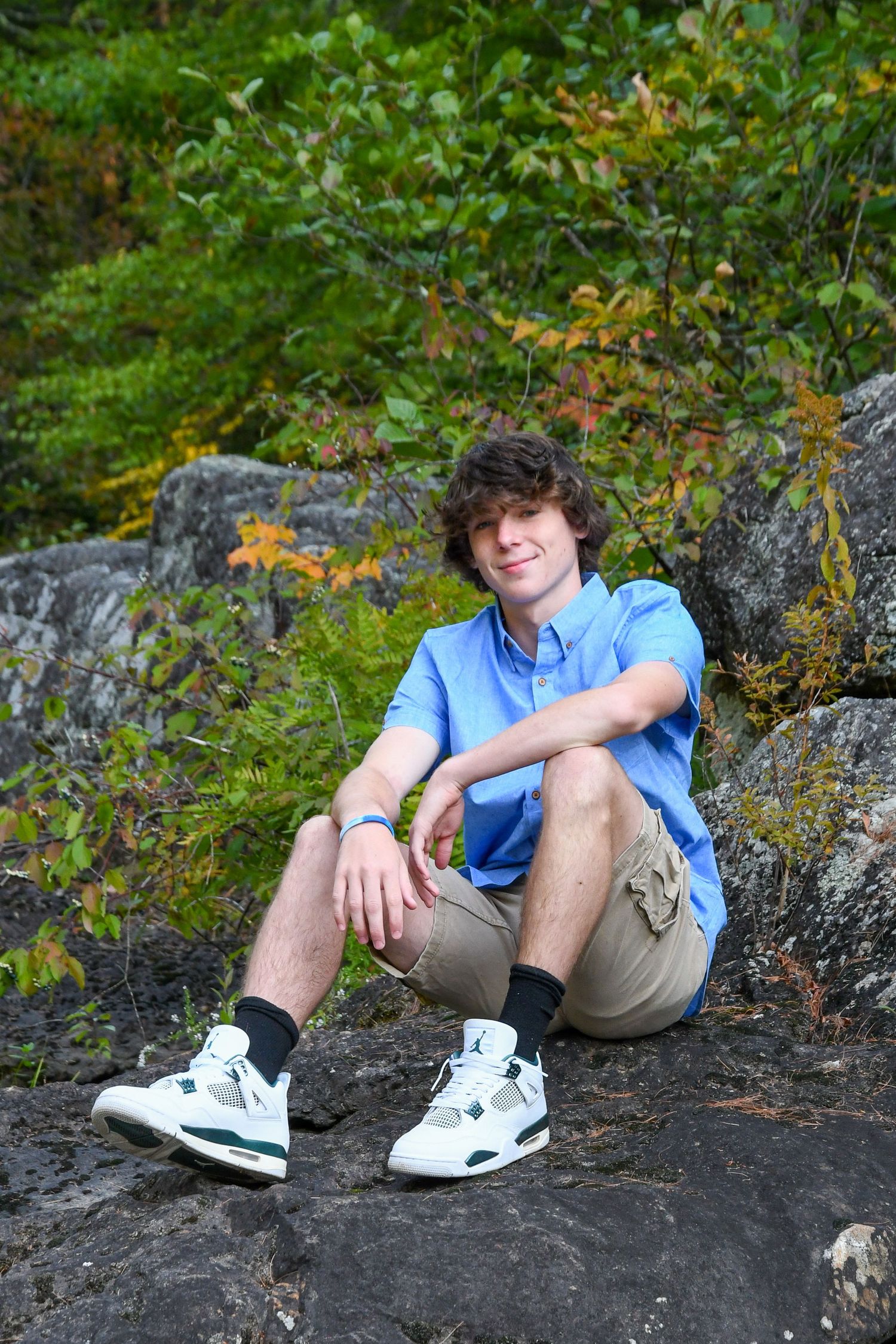 Person in blue shirt and khaki shorts relaxes on rocky ledge with green forest background.