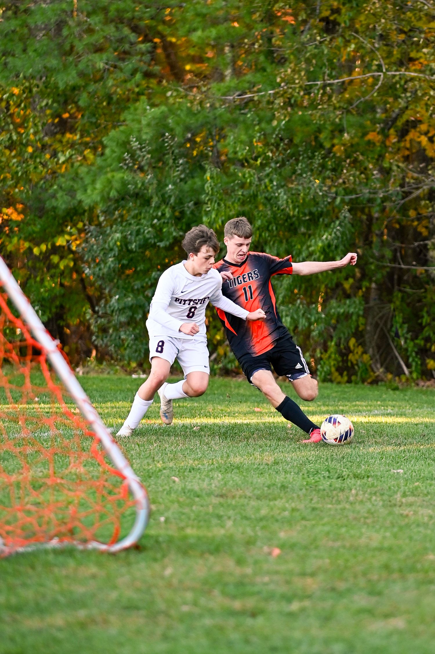 Two soccer players compete for the ball during a match on a grass field with autumn foliage in the background.
