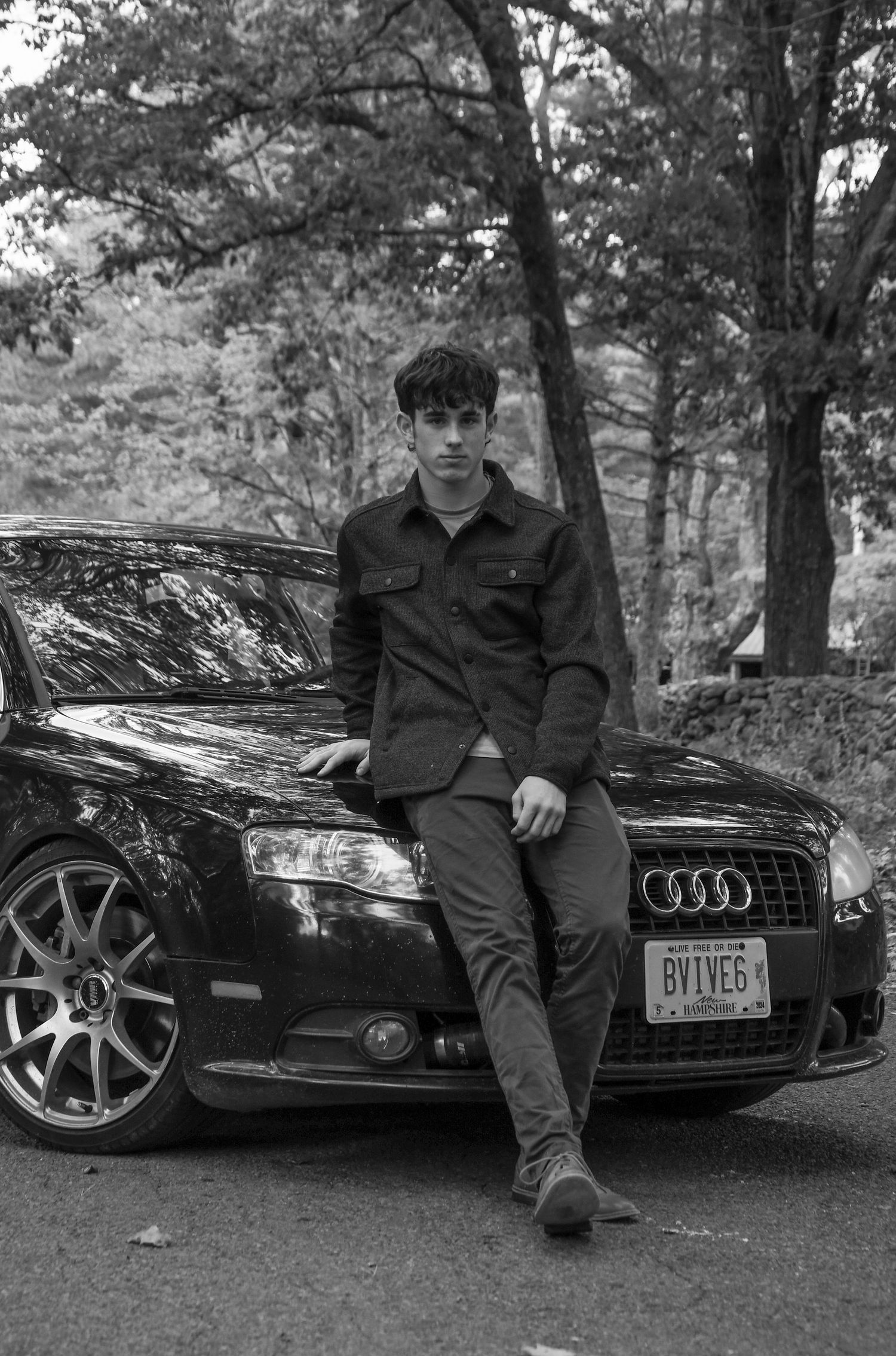 Black and white photo of person posing with Audi sedan on forest road.