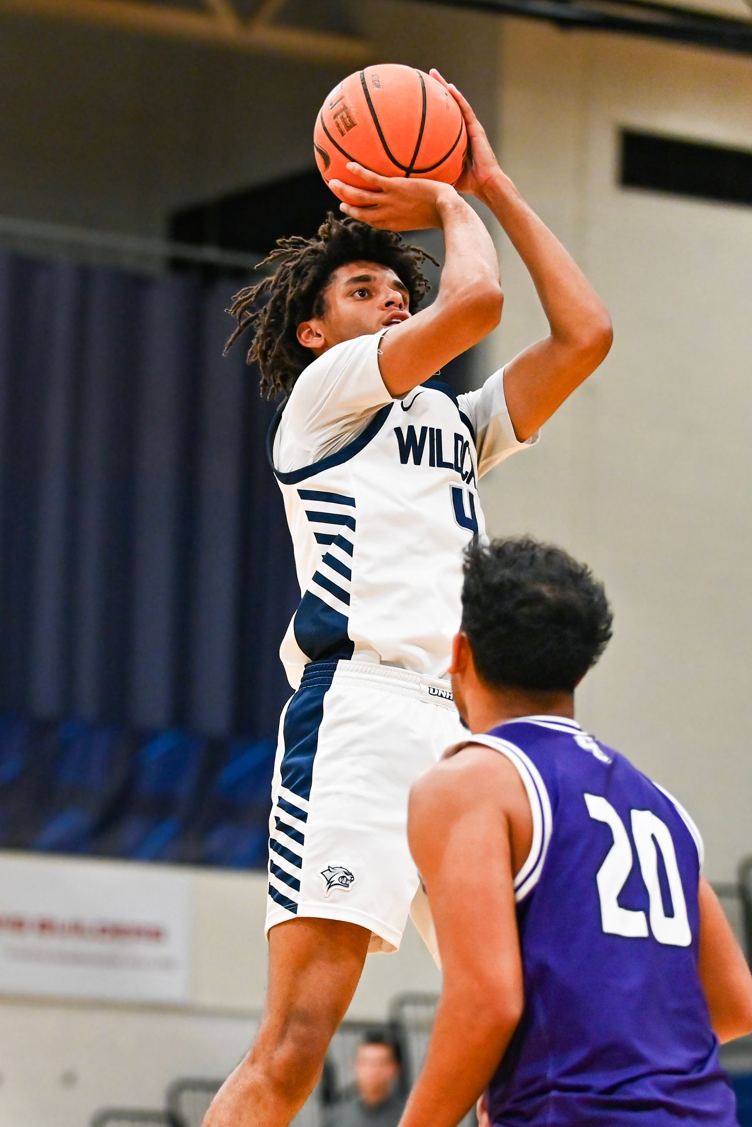 Basketball player in white jersey shoots over defender in purple jersey during competitive game action.