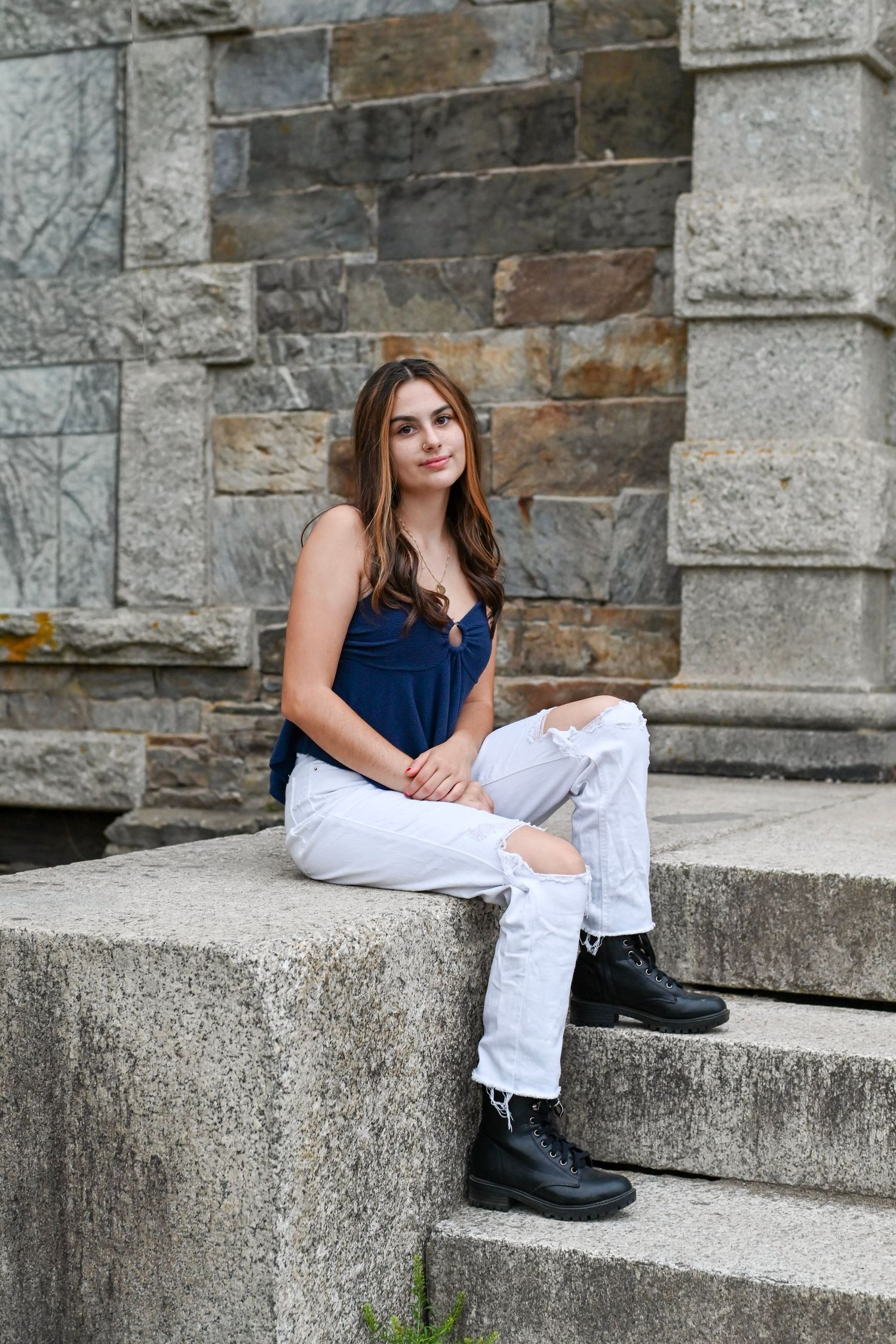 Person in navy top and white pants sits casually on stone steps against rustic stone wall background.