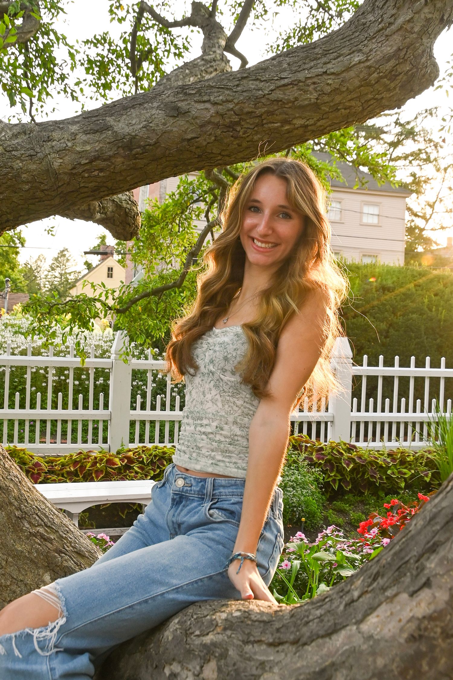 A person relaxes on a large tree branch at sunset with a white picket fence and garden in the background.